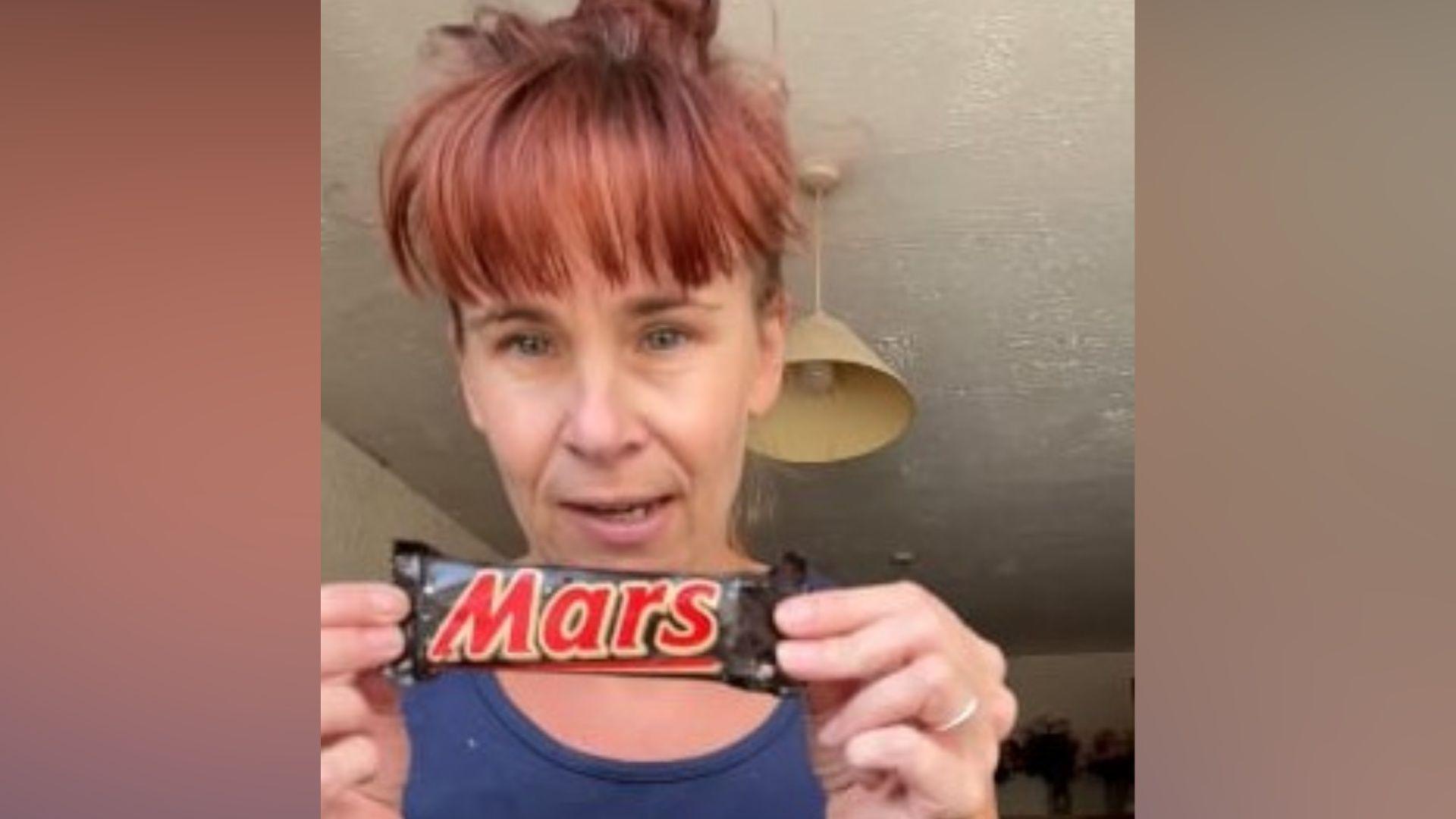 A woman holding up the old Mars Bar. She is wearing a navy vest and has red hair tied up with a full fringe.