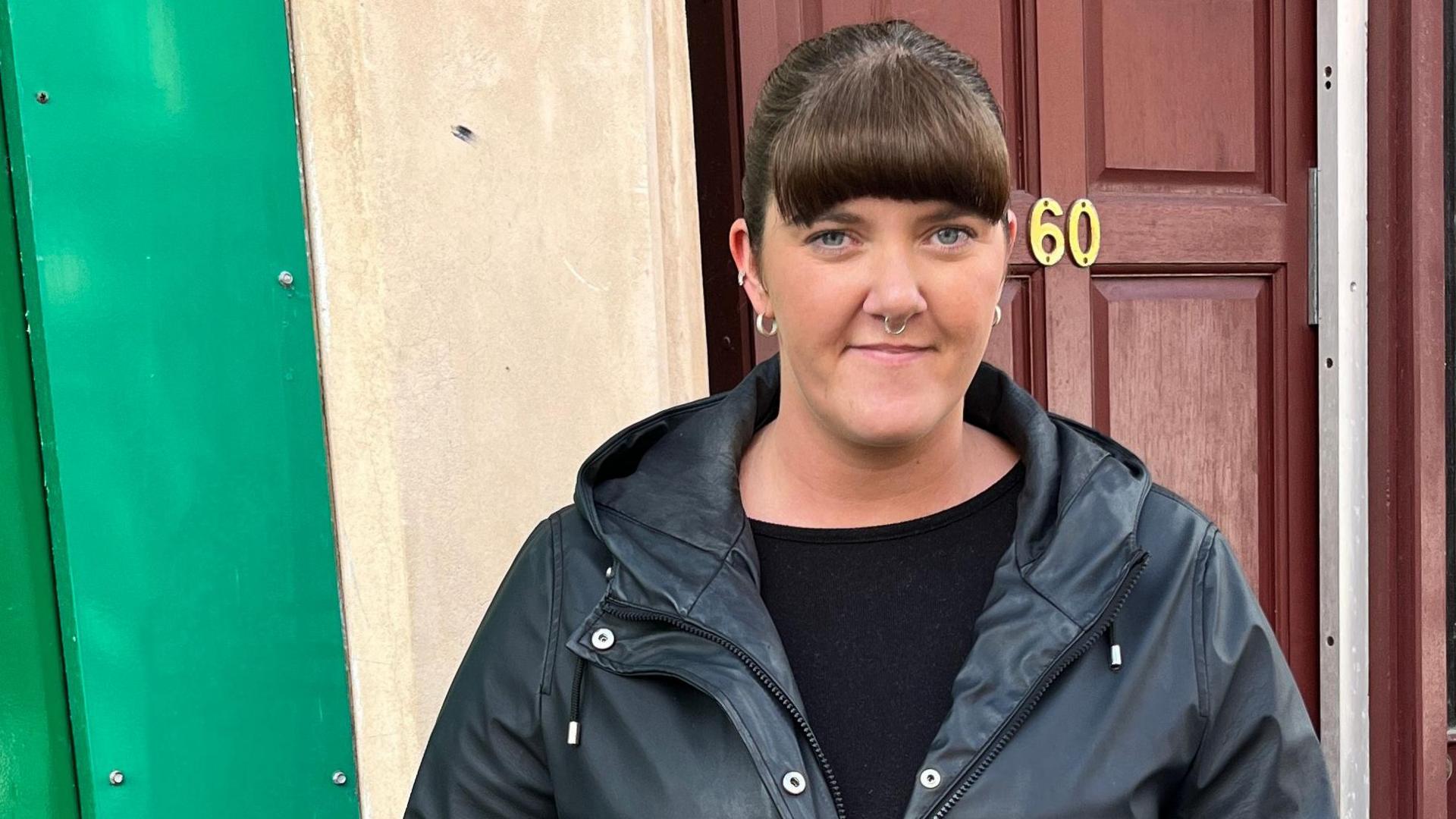 Nicole Copland stands in front of a brown wooden door, wearing a black top and jacket. She has brown hair which is tied back.