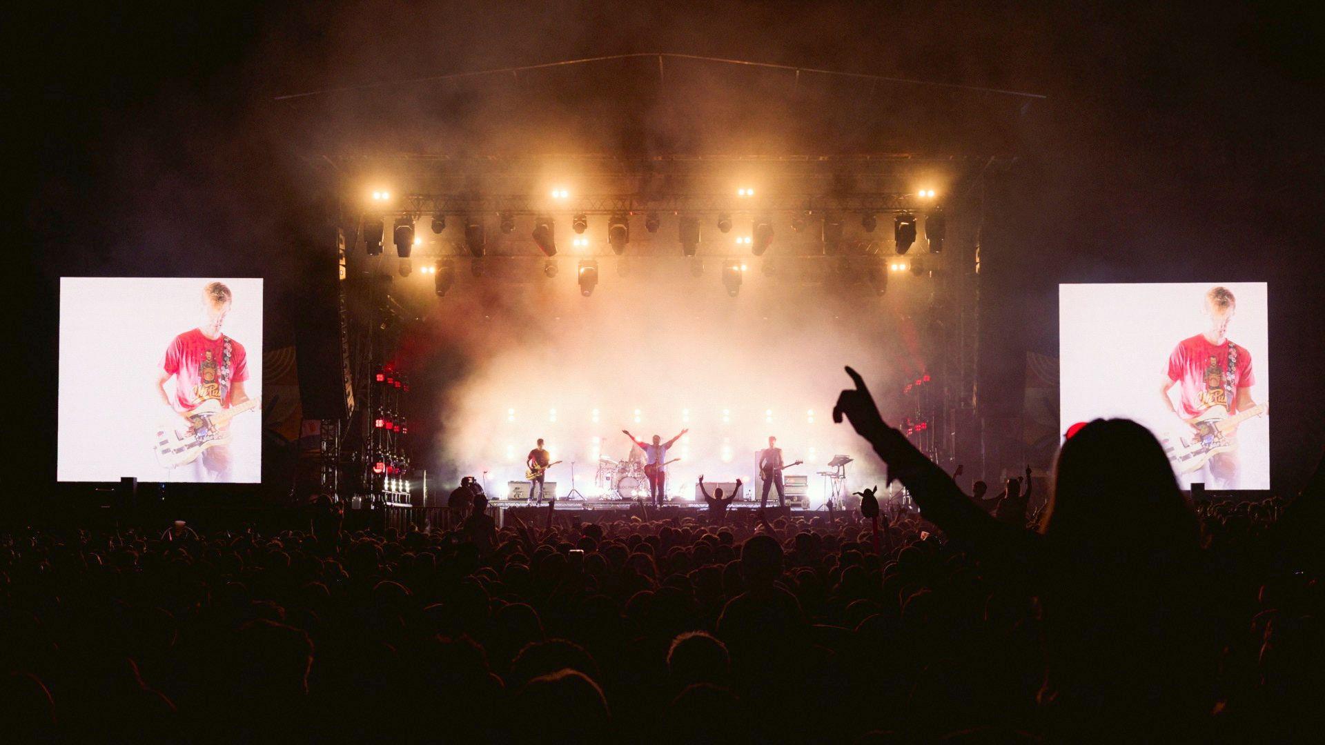 Bloc Party is performing on stage. It's dark apart from the stage which is lit up using golden lighting and there are two large screens either side showing the performers close up. The picture is taken from the audience and shows people with their hands raised.