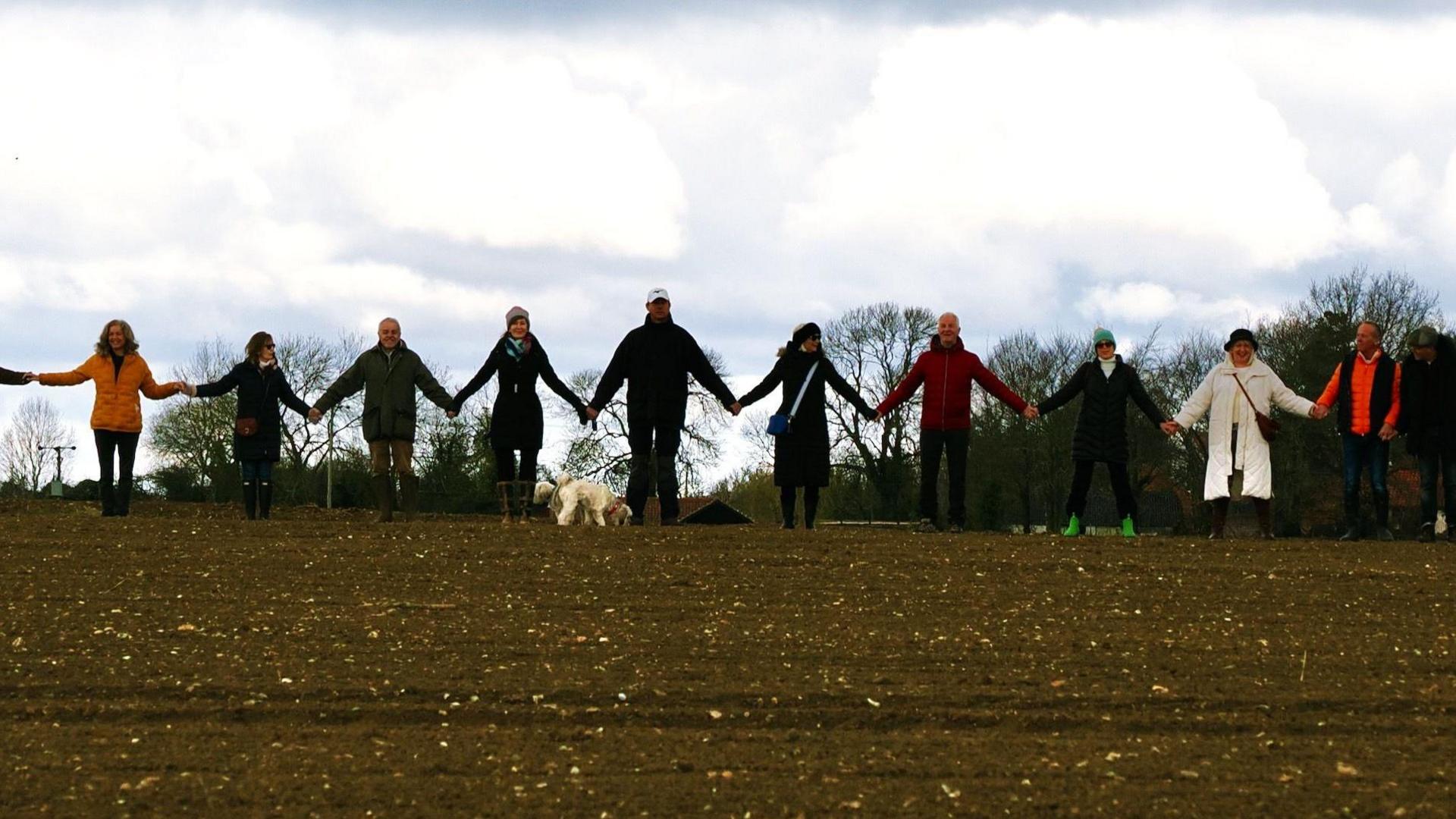 About 10 people hold hands in a ploughed field. A dog is standing in front of them.