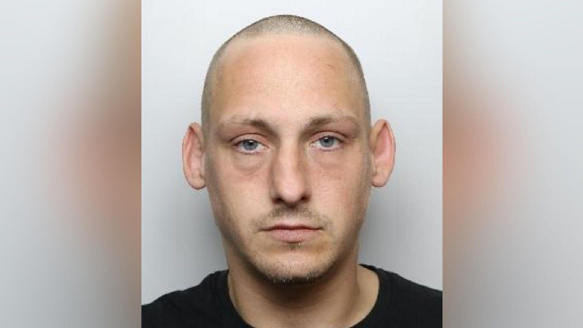 A mugshot of Corney, who has a shaven head and a little facial hair above his upper lip and along his chin. He is wearing a black top and looks straight at the camera