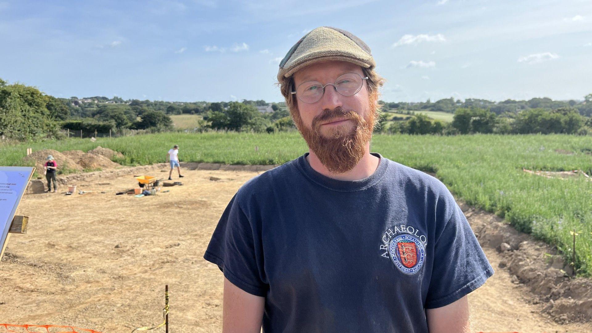 Dr Hervé Duval-Gatigno, with a beard and wearing a dark blue T-shirt and a flat cap, standing in front of archaeological dig