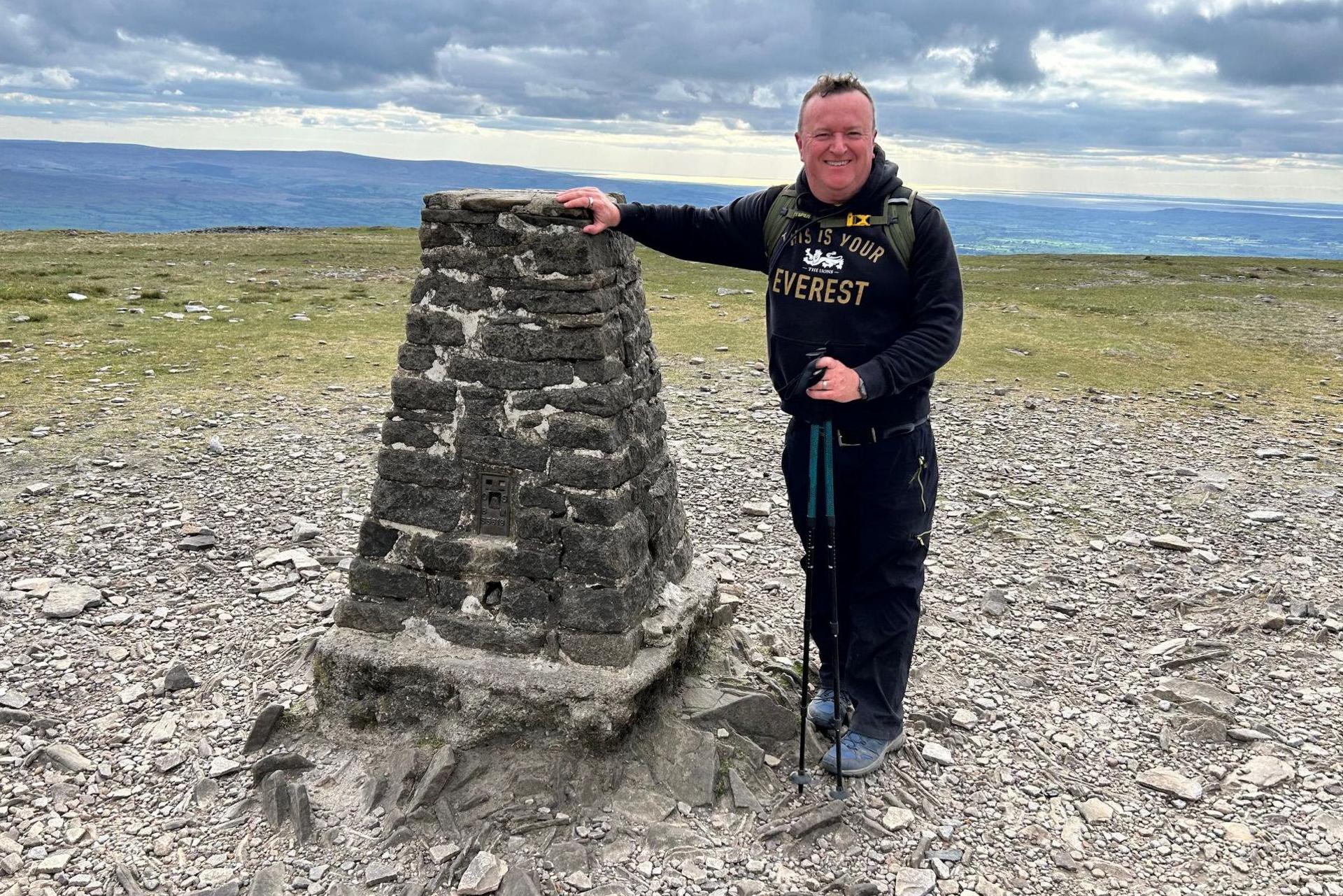 Mark Singleton wearing a black top and black casual bottoms and trainers. He is smiling and holding two walking sticks. He has short dark hair and is leaning against a stone mound on top of a mountain peak with rocks and grass. There are mountains and a cloudy sky behind.