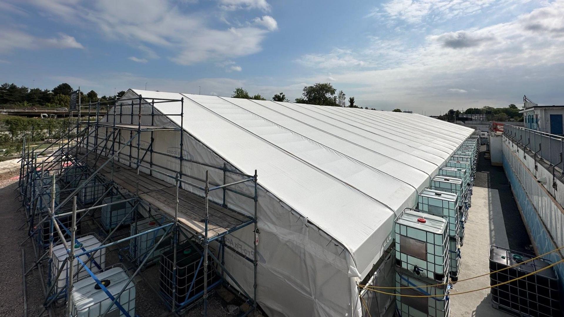 Marquee-style tent covered in white plastic with scaffolding at the nearest end and two-tier water tanks placed along its sides.