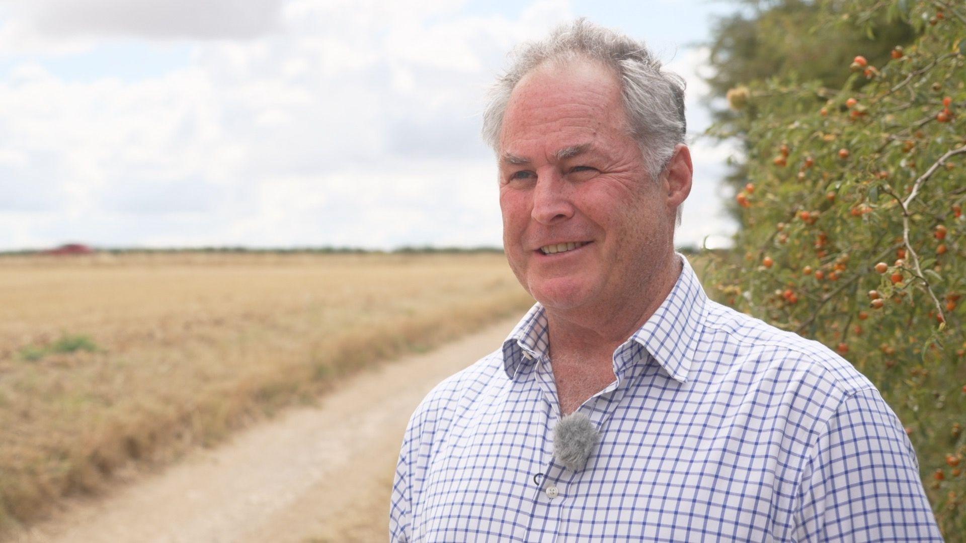 A balding grey haired man smiles while standing in front of a hedgerow laden with red berries. To the left we can see a field of barley recently harvested.