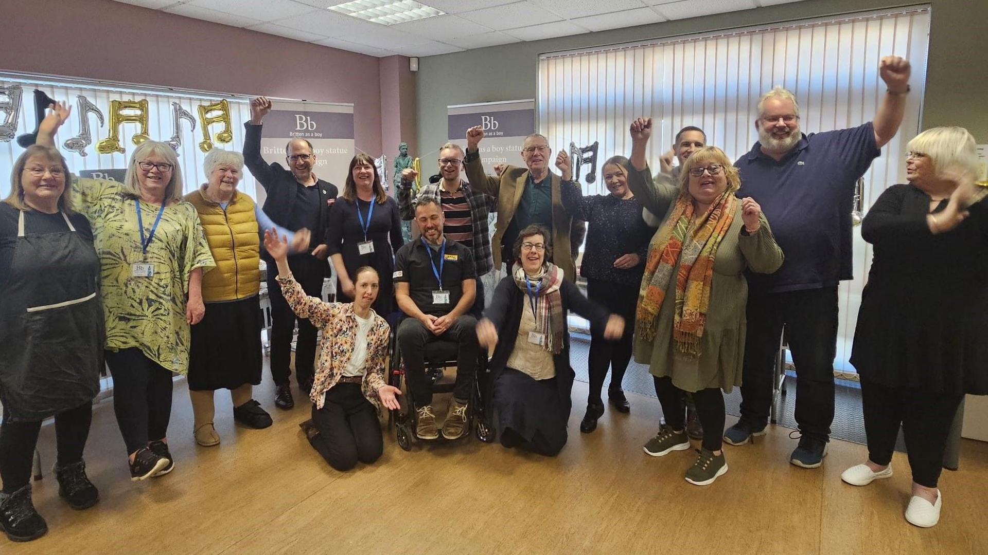 In a room with a wooden floor, fifteen people, including a man in a wheelchair, raise their arms to celebrate the target being reached