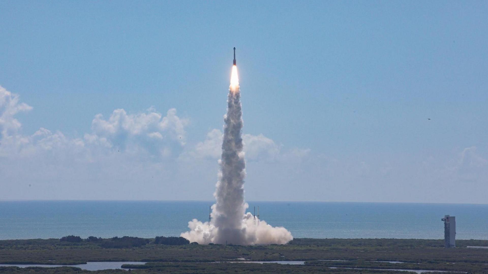 A rocket takes off vertically  into a clear blue sky with the dark blue sea in the background. White-yellow blazing rocket fuel comes directly out of the rocket and then changes to a thick plume of white smoke.