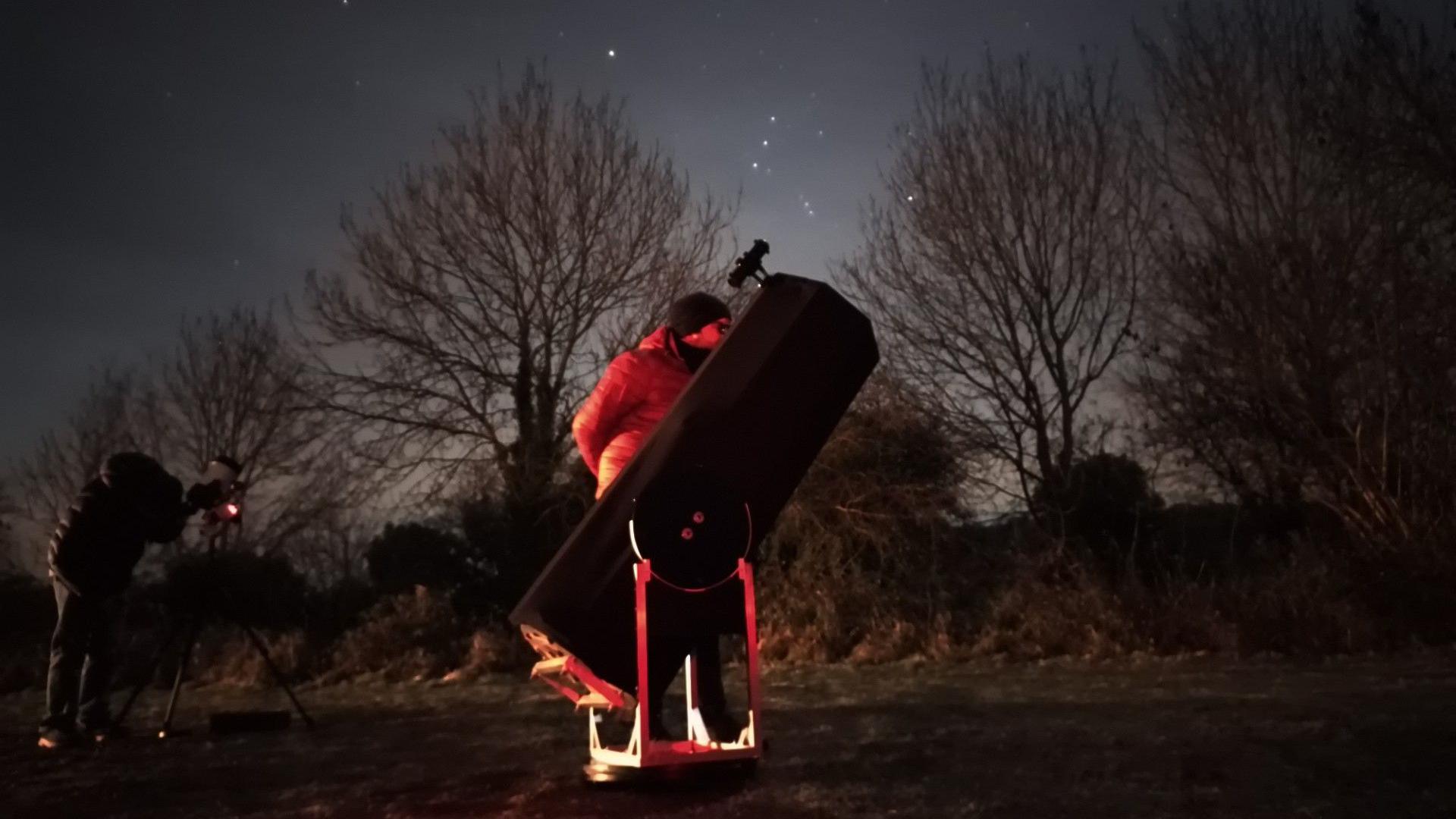 Ben Luger observing at Wellow village with Bath Astronomers. He is looking through a professional grade telescope and is standing on the right side of it, wearing a black beanie hat and thick clothing. The trees are silhouetted against a starlit sky.