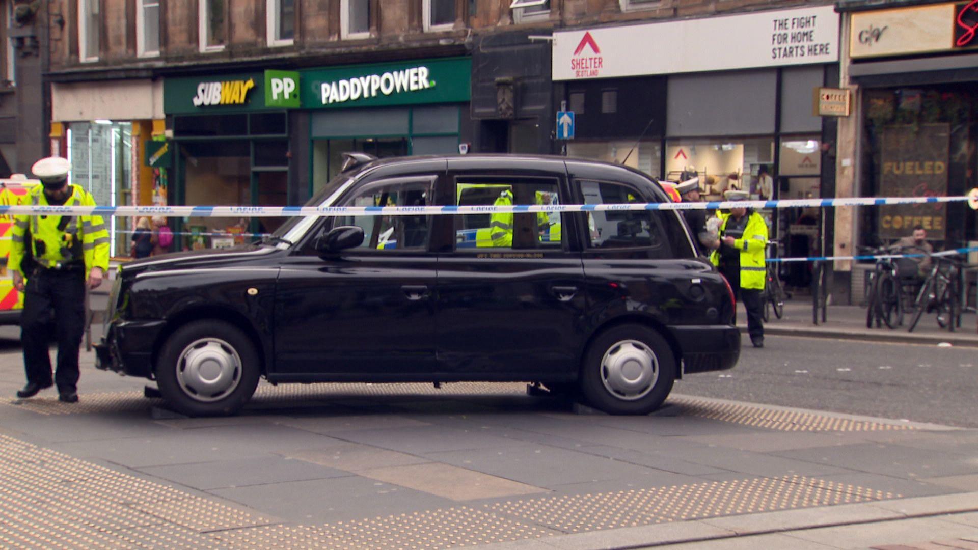 Police officers in yellow coats stand beside a black cab  behind police tape