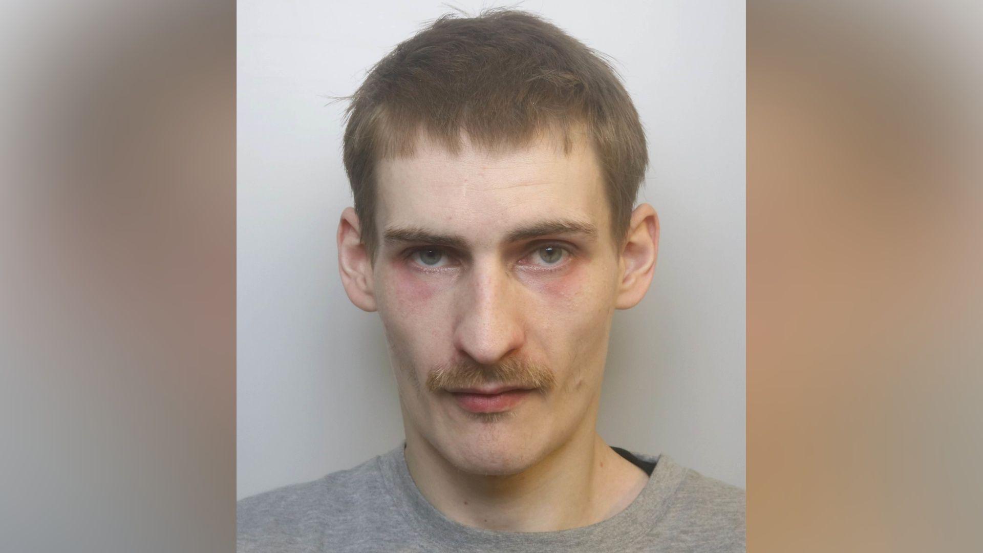 A mugshot of Gunter. He is looking at the camera with a neutral expression. He is wearing a grey jumper, and has a mustache and short brown hair. He has red marks under his eyes.