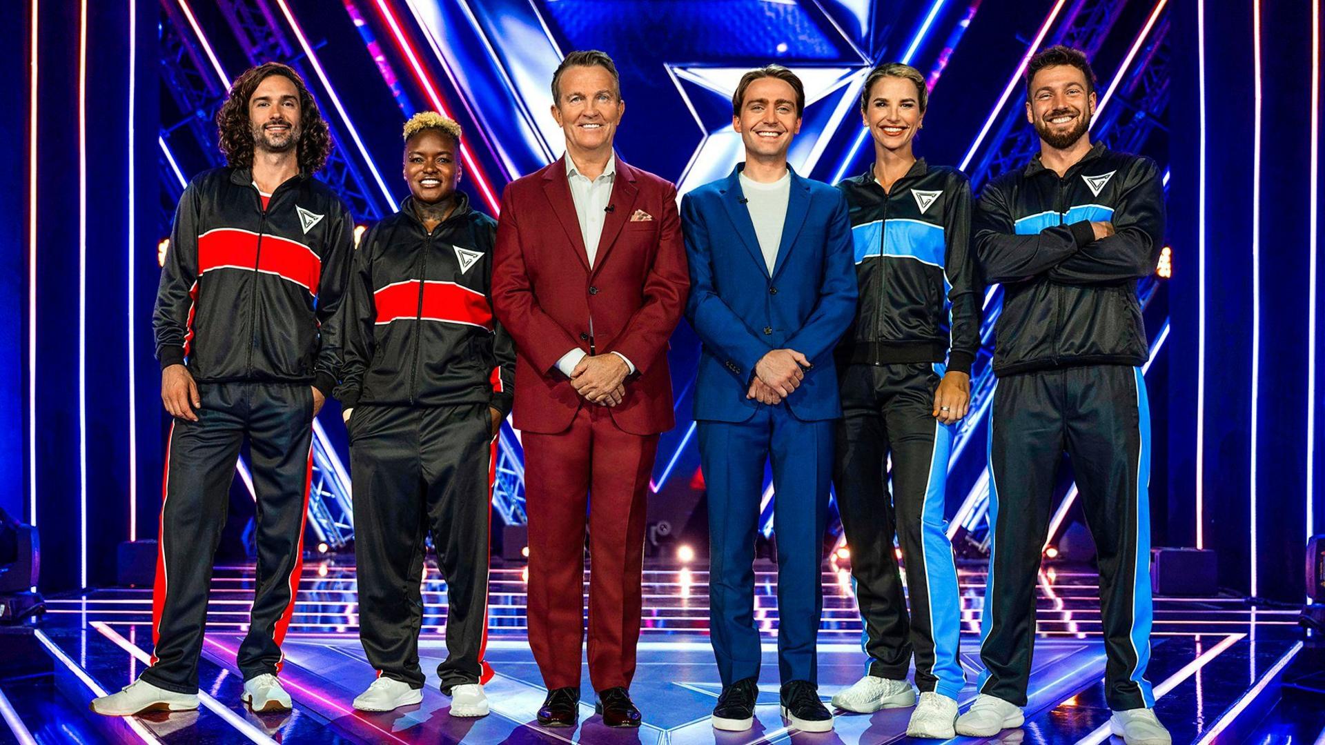 Four celebrities in black overalls stand next to the hosts of the Gladiators gameshow on a shiny floor studio.