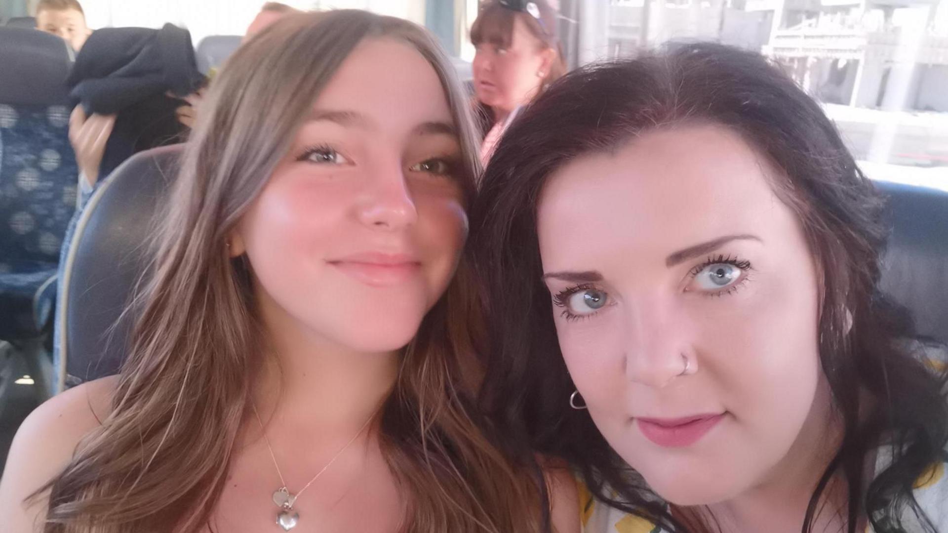 Chloe Longster (left) smiles with her mother Louise Longster (right). Chloe has long brown hair that has been curled, she is wearing natural makeup and has a silver locket around her neck. Her mother is looking at the camera and has dark brown hair, light red lipstick and a pierced nose. They are sitting on a coach together.