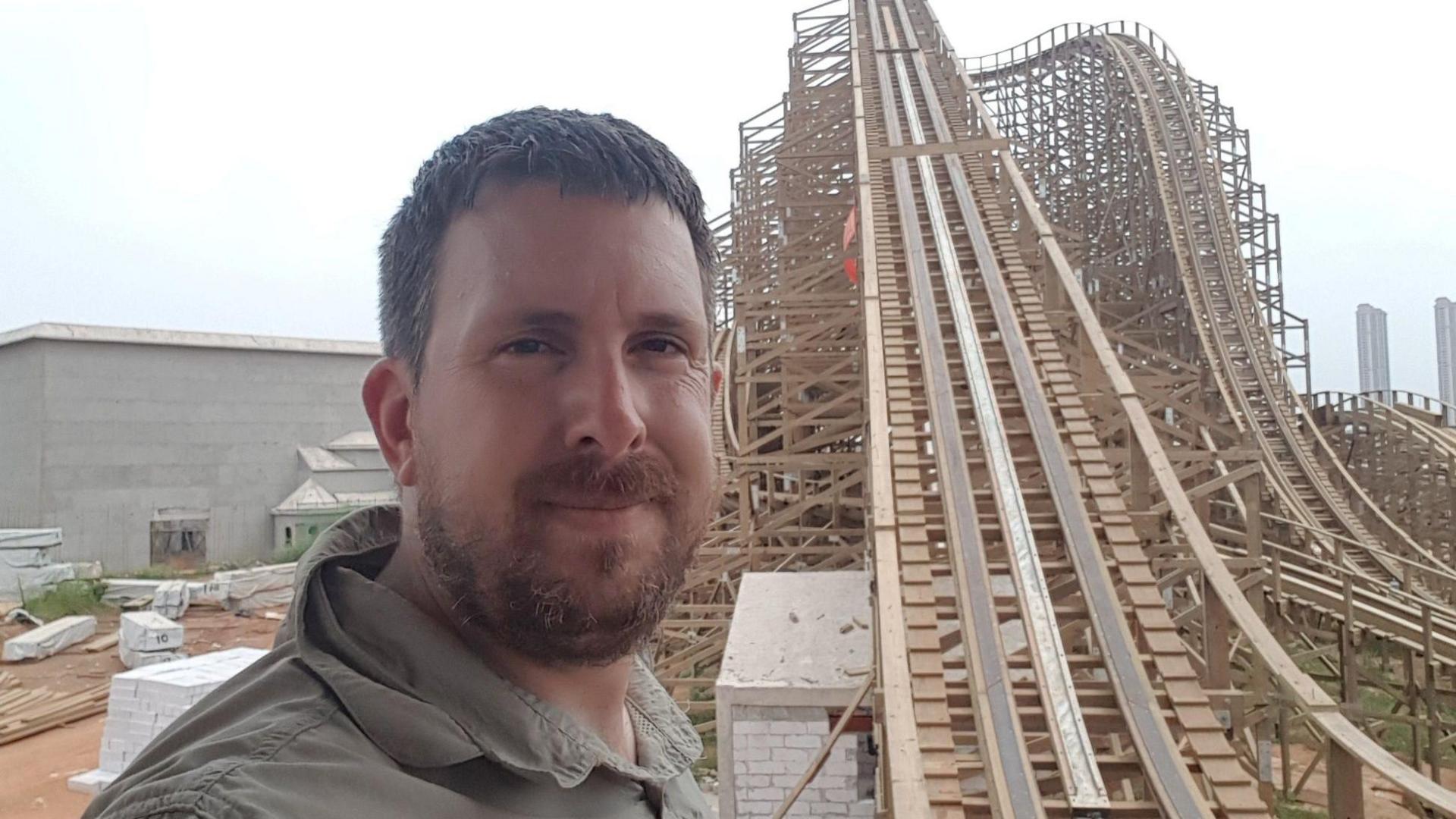 A man is stood at the bottom of a wooden rollercoaster, the track of which has a steep incline. He has a beard and is wearing a green top.
