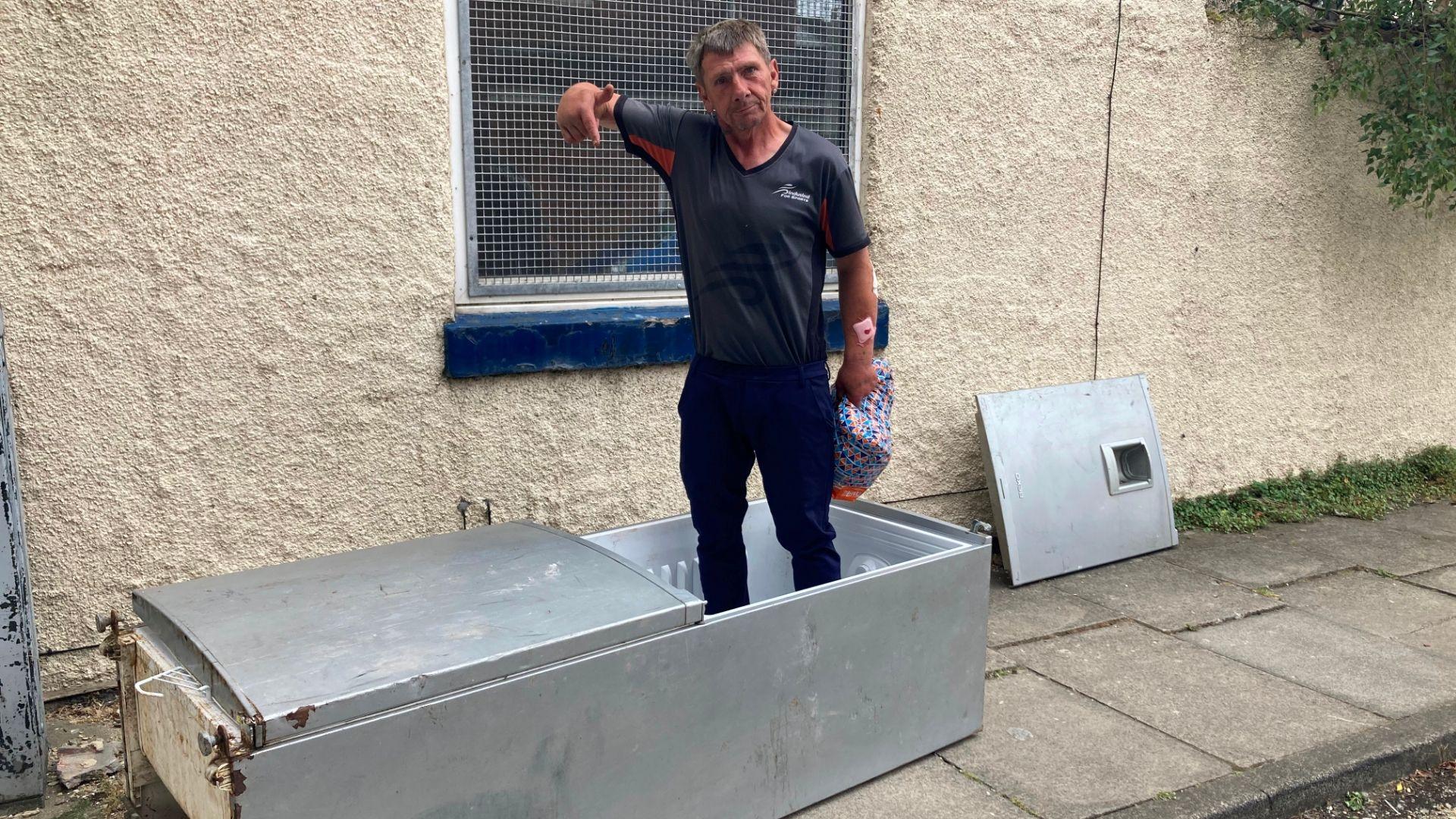 A man in a grey top stands in a fly-tipped fridge pointing at it.