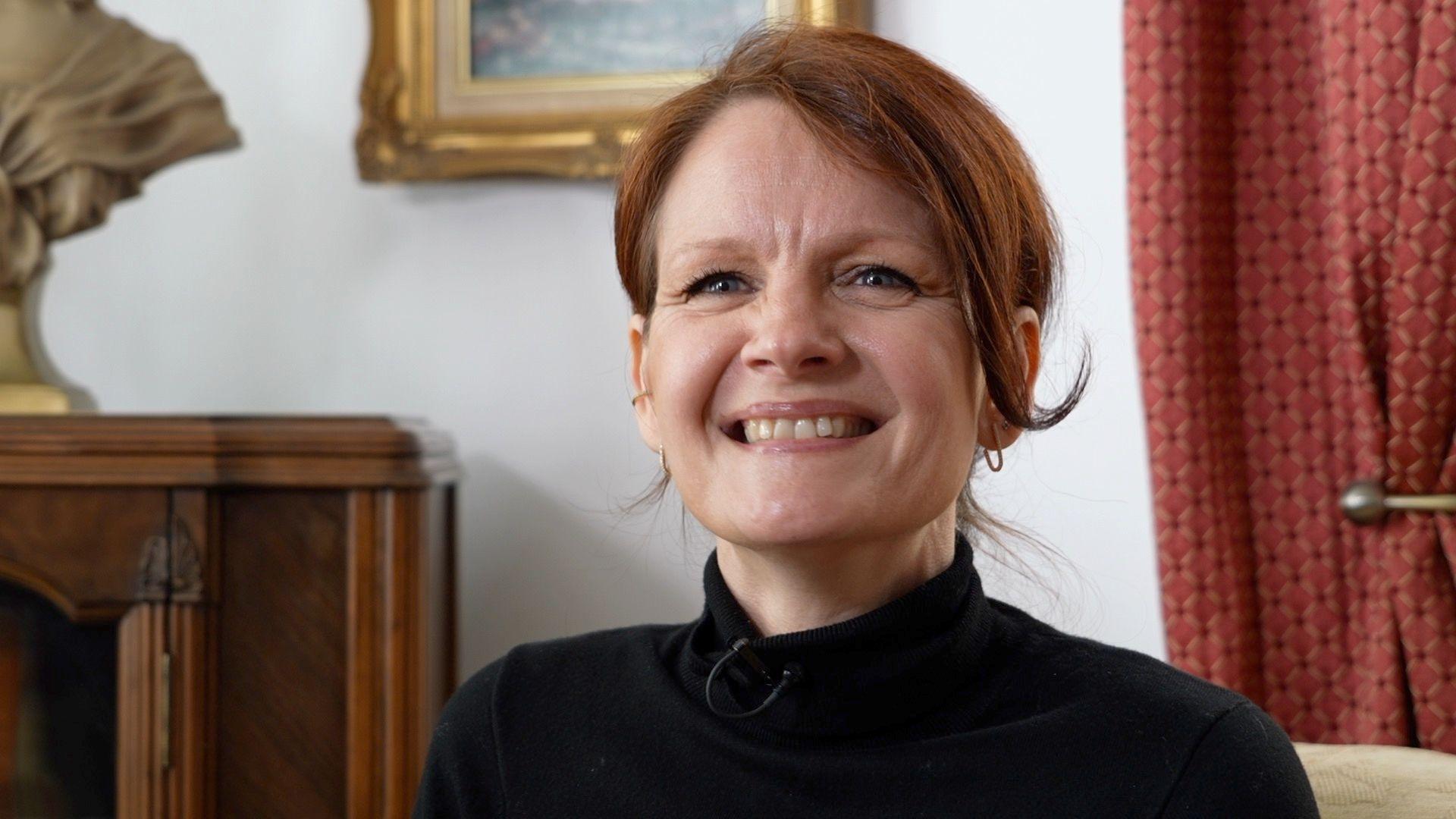 A woman with red hair tied back is sitting down in a sitting room, with a wooden unit to the left of the image and a red curtain tied with a brass tie back behind her. An ornate gold picture frame is on the wall behind her. She wears a black polo neck top and is smiling.