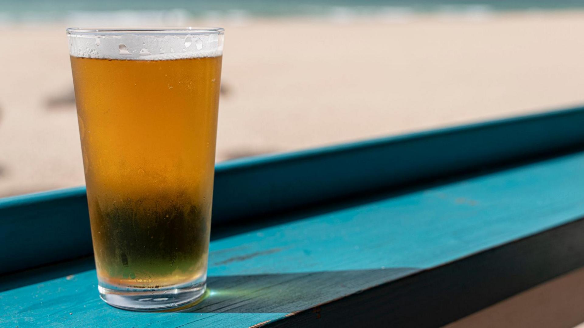 A generic picture of a glass of lager on a green piece of wood, with a beach and the sea in the distance.