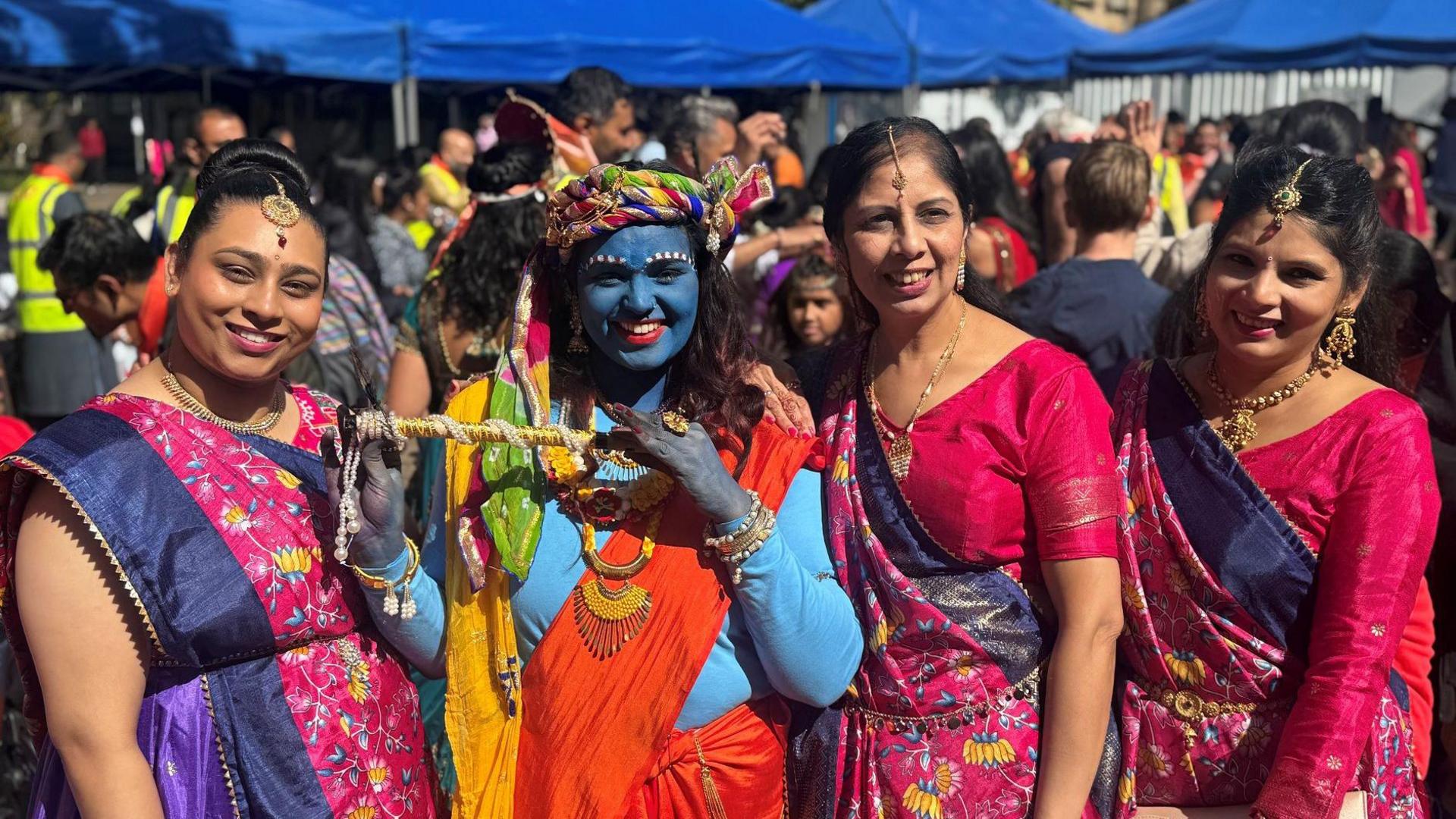 Group picture of four Hindu women smiling at the event. One of them is wearing blue facepaint and top with an orange toga draped over a shoulder as she depicts the Hindu deity Lord Krishna. She holds up a flute and wears a multi-coloured headwrap plus gold-coloured necklaces and bangles. The three ladies around her wear identical blue, pink and purple saris.
