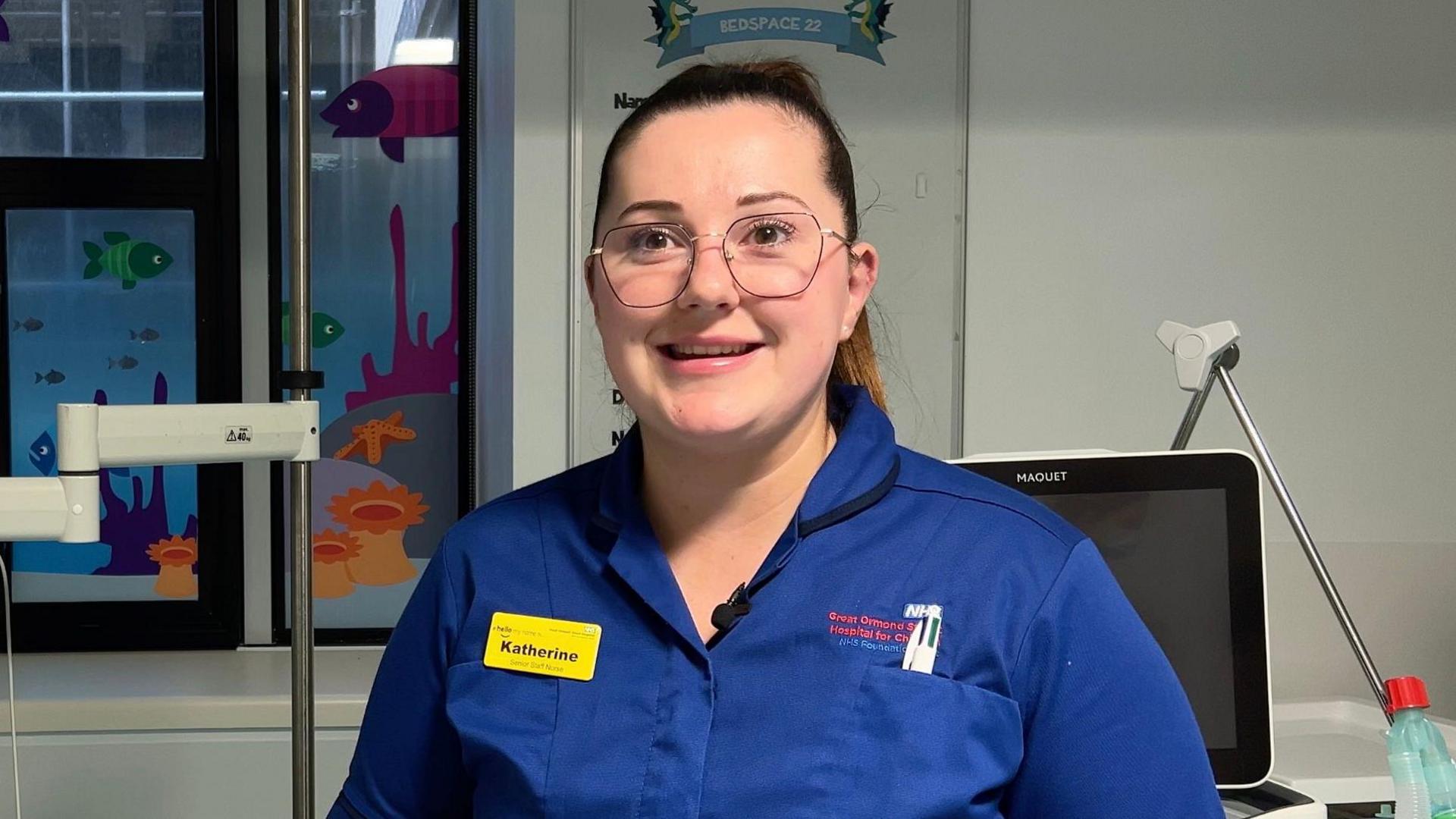 Nurse Katherine, wearing a blue uniform and glasses, smiles while surrounded by medical equipment and colourful decorations of fish on the windows.