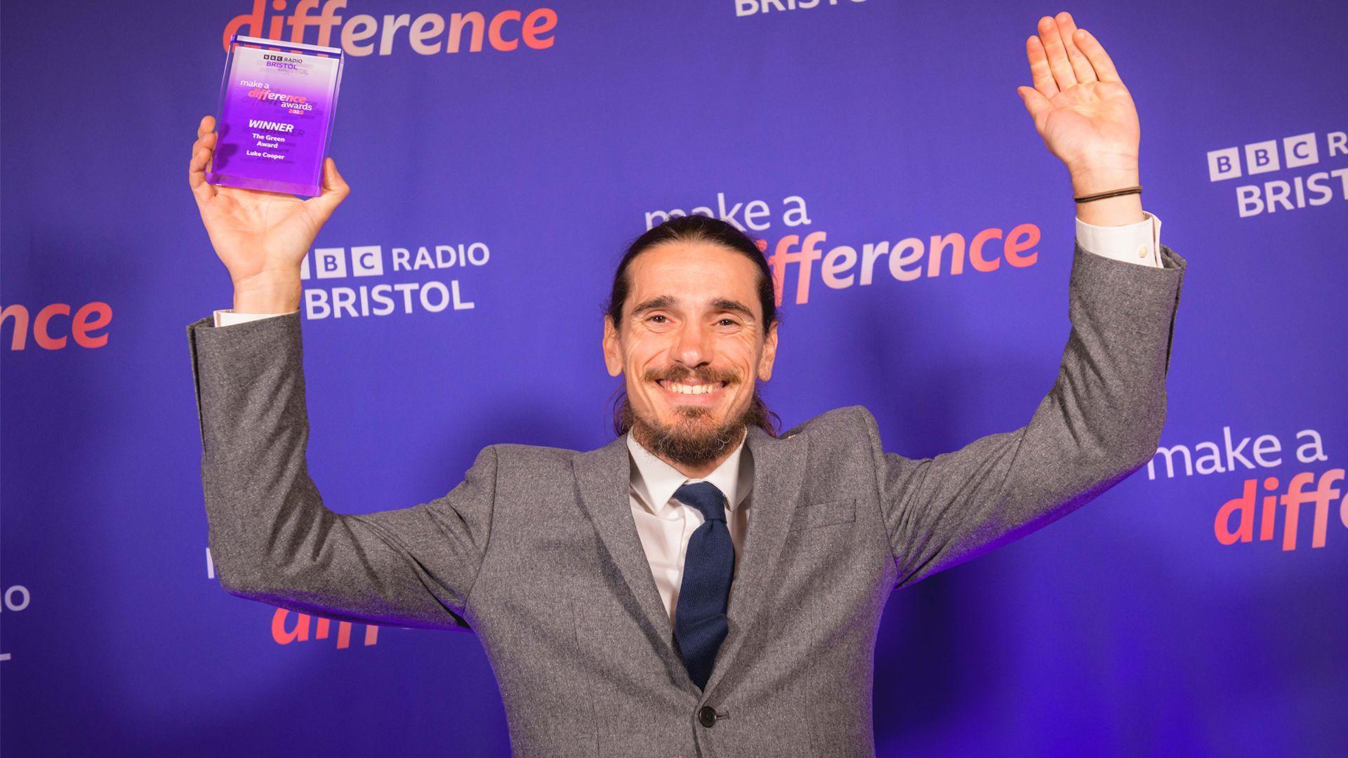 A man wearing grey suit holds a purple trophy aloft whilst smiling at the camera with both arms in the air. The background is purple.