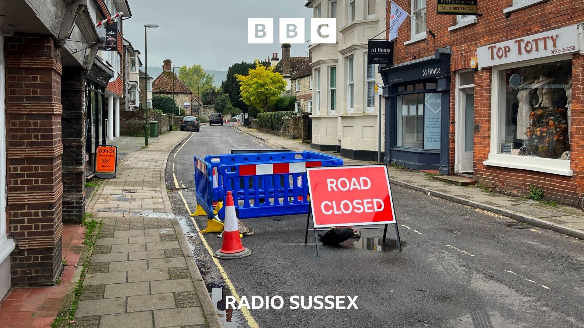 A small high street is partly closed by a sinkhole on one side. Blue plastic fencing surrounds the hole and a red ROAD CLOSED sits in the middle of the road.