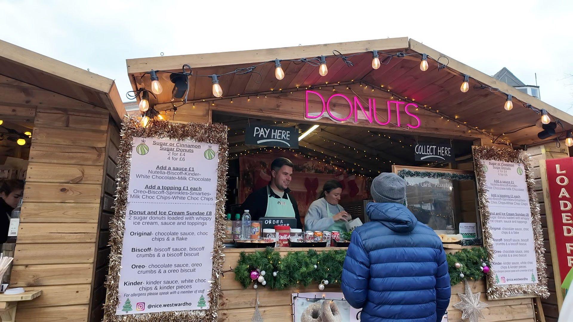 A wooden stall with "Donuts" written in neon pink lights above the hatch. There are orange Christmas lights hanging from the awning of the building. A man in a blue puffer coat is queuing to buy something from two stallholders. 