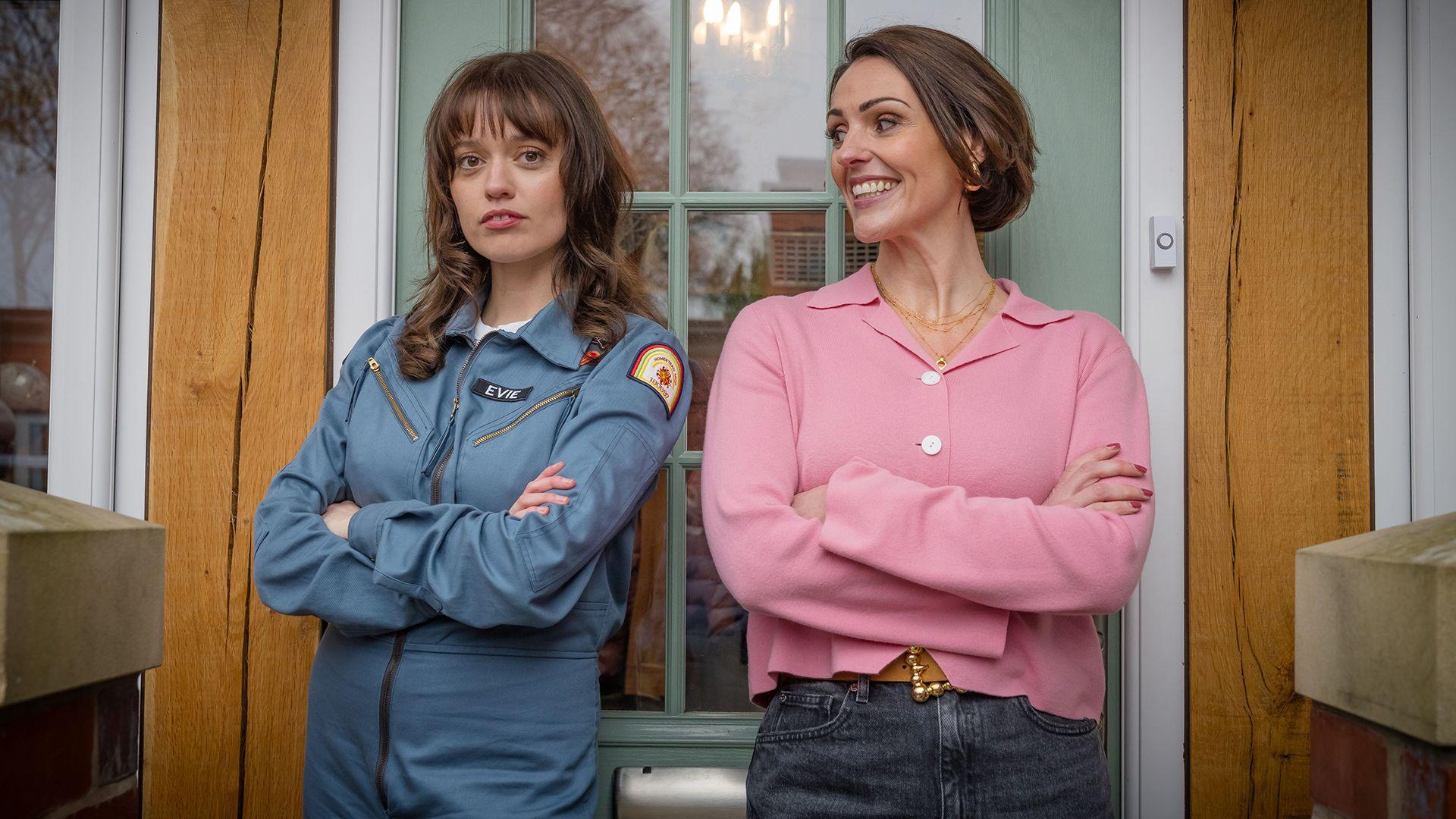 Aimee Lou Wood and Suranne Jones stand in front of a green front door, both with their arms crossed, dressed as characters Evie and Suz in a publicity still from Film Club. Wood looks directly at the camera with a straight face while Jones smiles and looks at Wood.