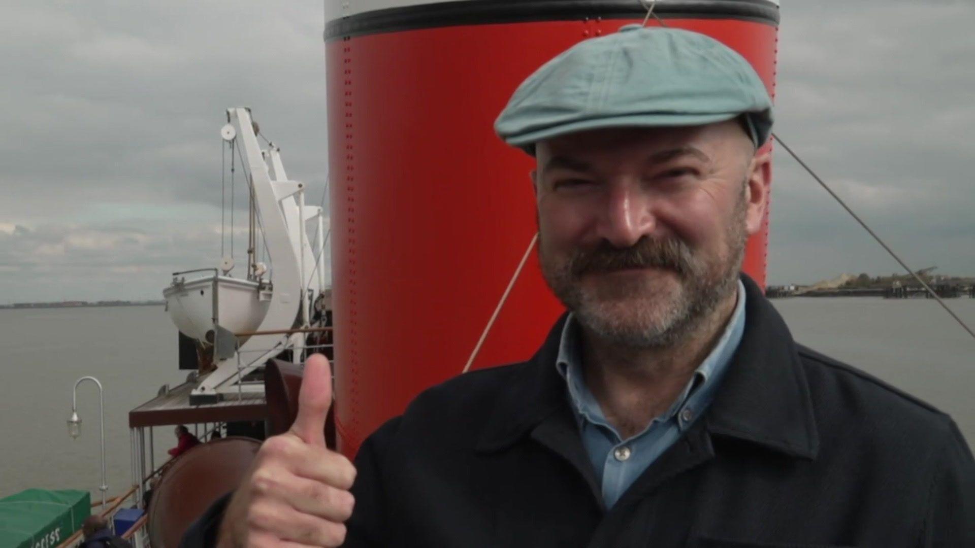 A man stood on the Waverley while it is at sea, with a large red chimney and lifeboat cranes directly behind him. He is wearing a hat, smiling, and giving a thumbs up to the camera.