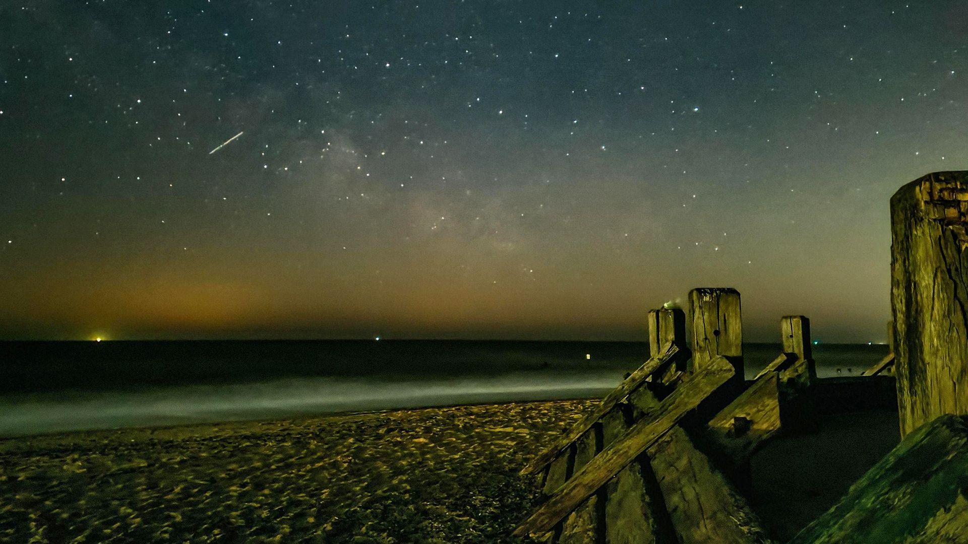 night sky image from a beach overlooking the sea. Lots of bright white spots in the sky and one streak of light showing a meteor streak