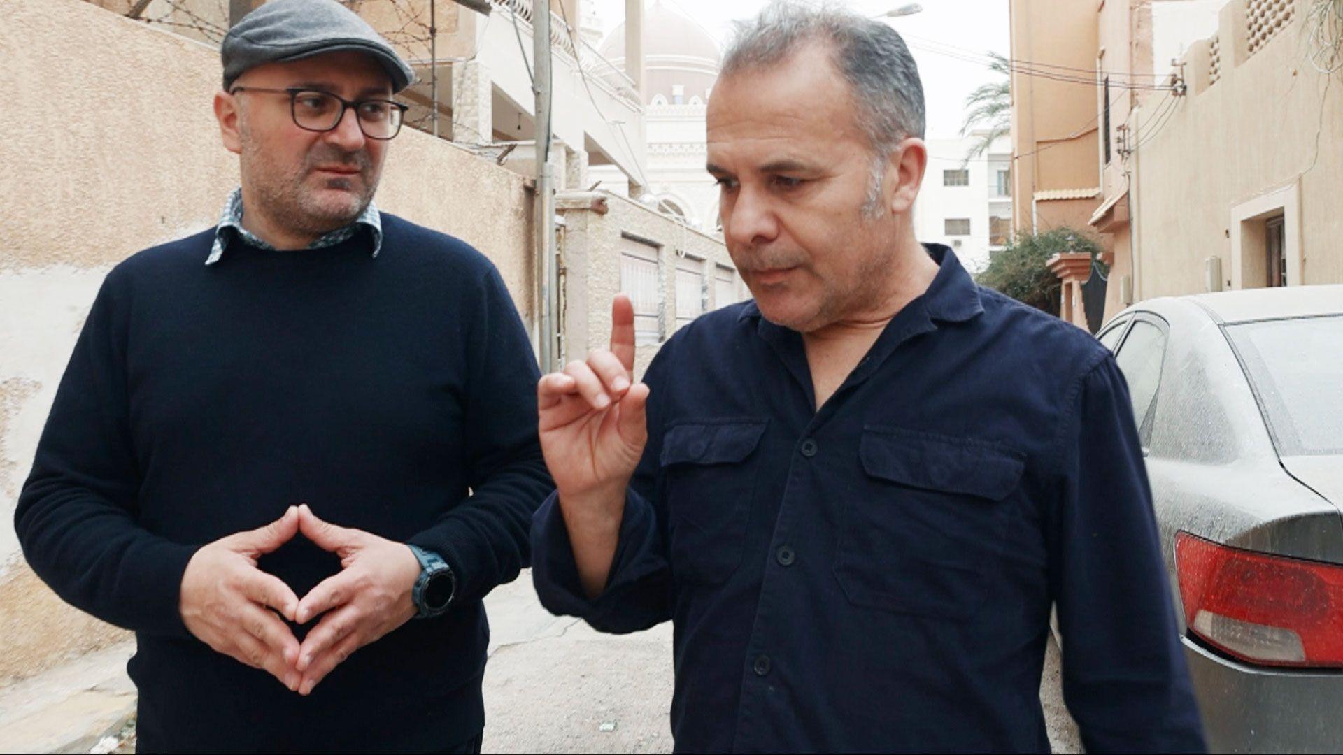 Kassem points a finger in thought as he talks to Moe steepling his fingers in the street in Tripoli. They are both wearing navy blue tops and Moe has glasses, a slight beard and a grey cap. The walls behind them are of a dusty sand colour.