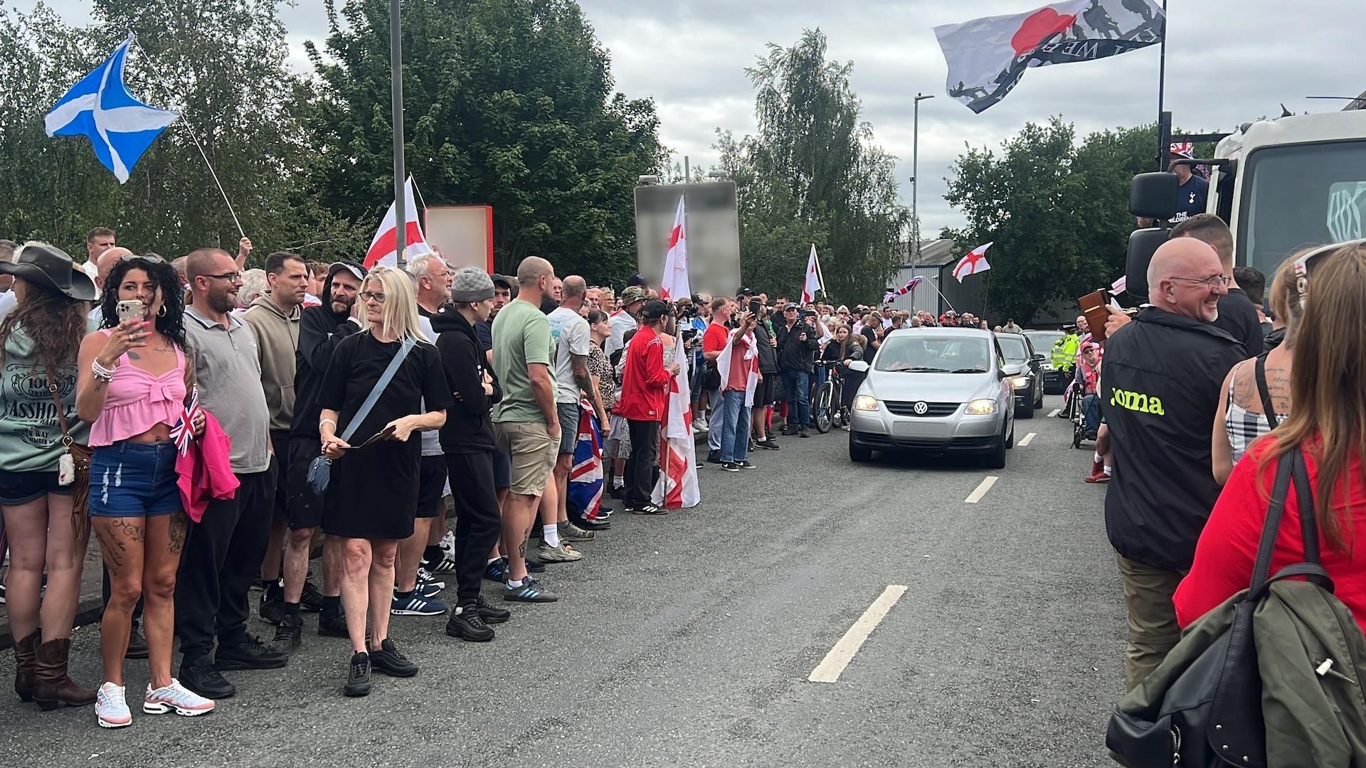 Scores of people standing on the road, many with St George's flags. A car is driving through the middle.