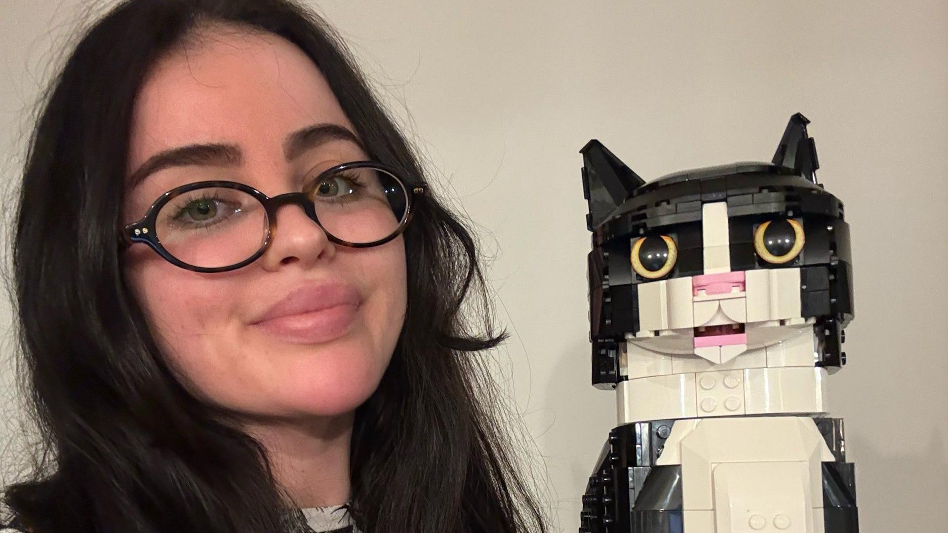 Elle Lynn with black-framed glasses and long dark hair in a selfie with a black and white Lego cat she made