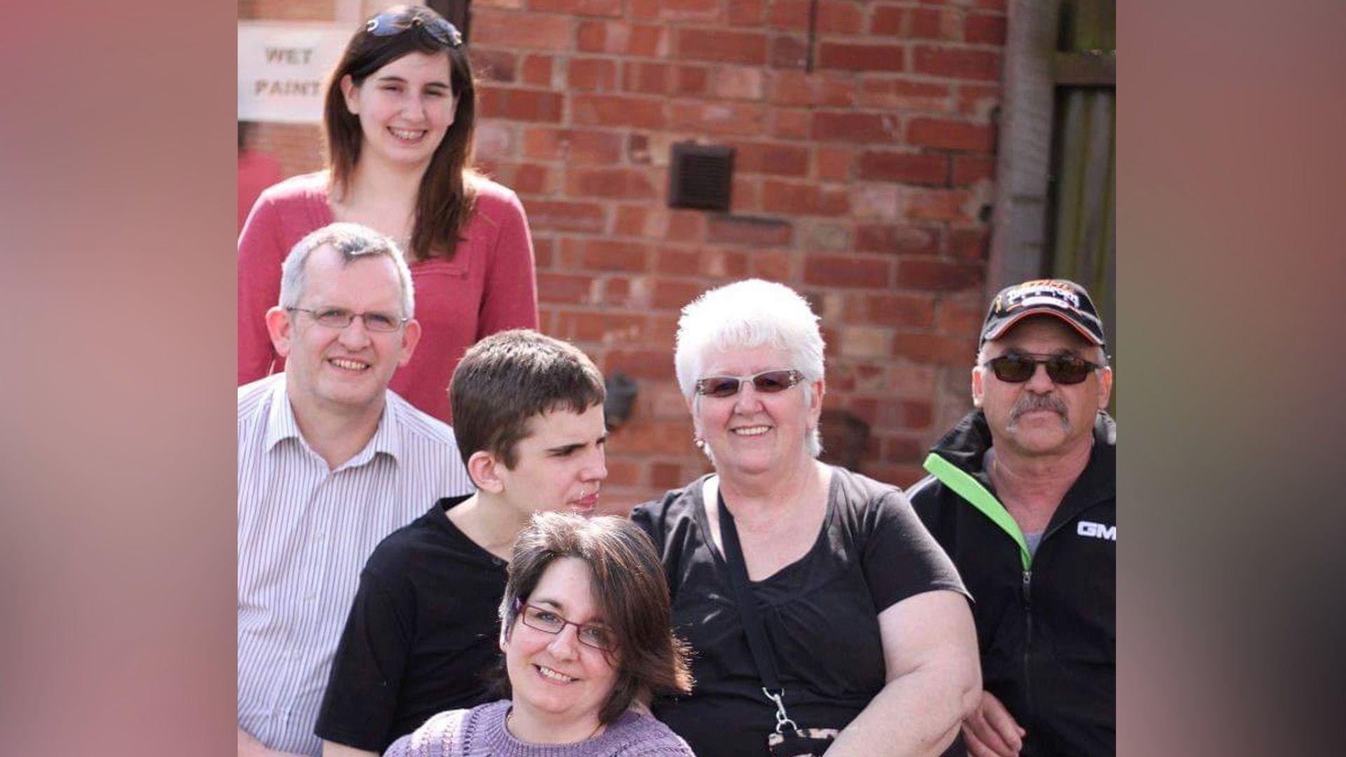 Declan with his family. His grandparents are also in the picture while Declan is looking to the side and wearing a black T-shirt. Declan's sister Kaitlyn has sunglasses on her head and is smiling, the rest of the family are also smiling, they've all positioned themselves around Declan.