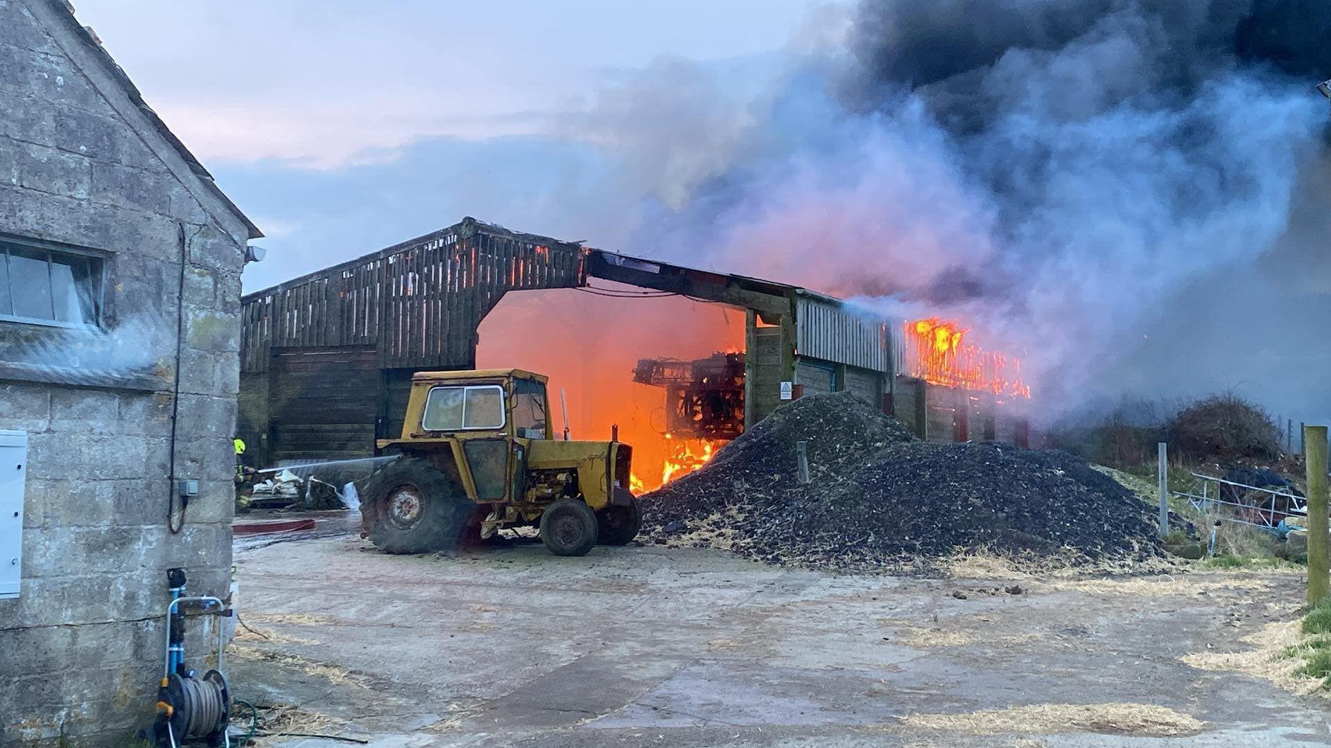 The large barn is well alight, full of flames. Black and grey smoke is billowing in to the air. There is a tractor parked near the barn door with two piles of aggregate next to it. The corner of a stone building is in the foreground.  