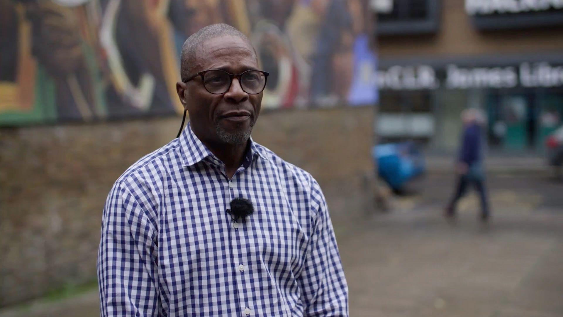 Peter Merrifield is wearing a checked purple and white shirt. The background is blurred and he is standing in front of a wall with graffiti in the background.