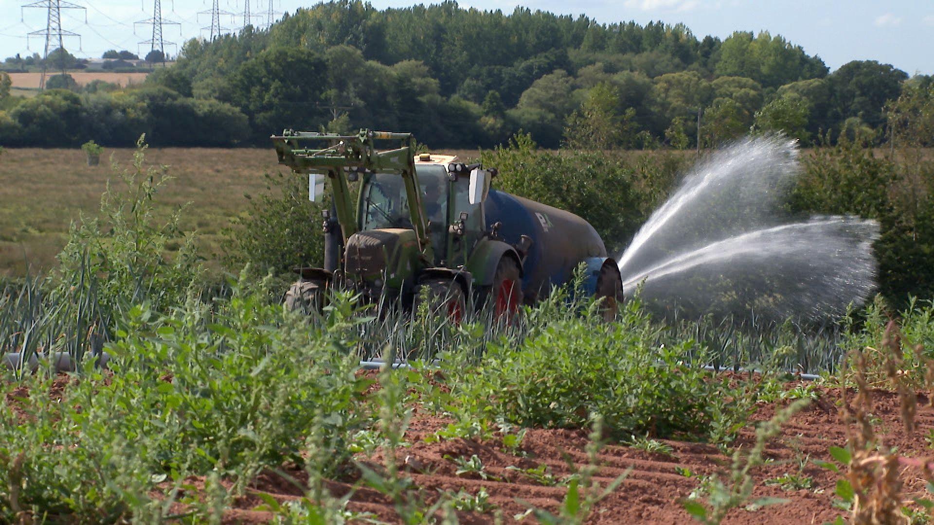 A green tractor, towing a large blue vacuum tanker, with water spraying from out of the back is working its way across a field of leaks