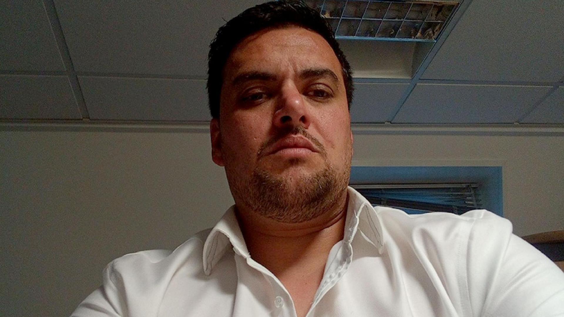 A head and shoulders image of Dominic D'Sa. He has short, dark hair and goatee-style stubble and is wearing an open-necked white shirt.
