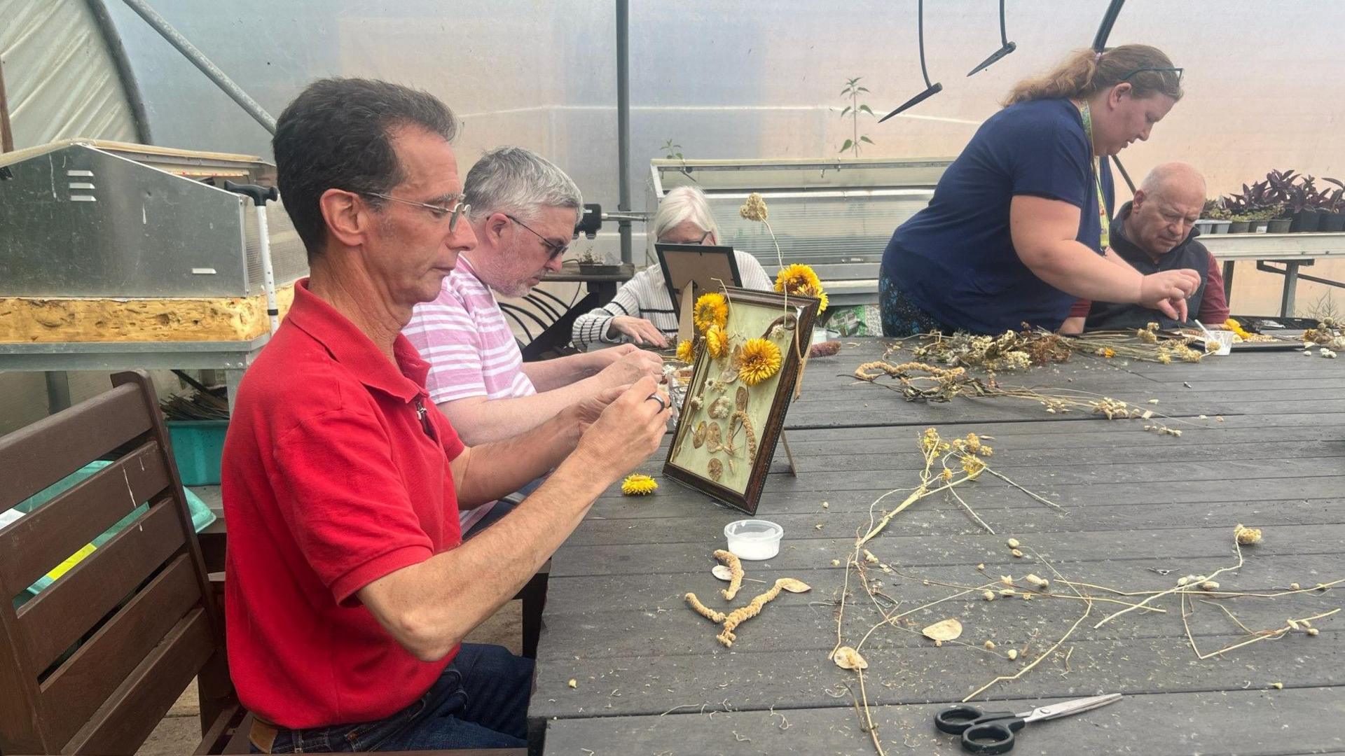 Four people sit or stand around a table in a greenhouse, making artwork out of flowers.