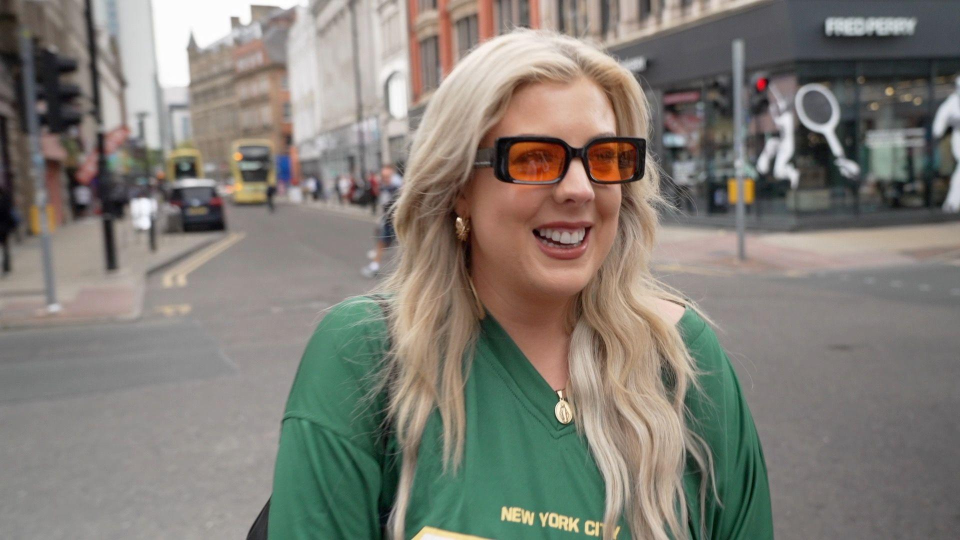 A smiling woman, with long blonde wavy hair. She is wearing orange sunglasses.