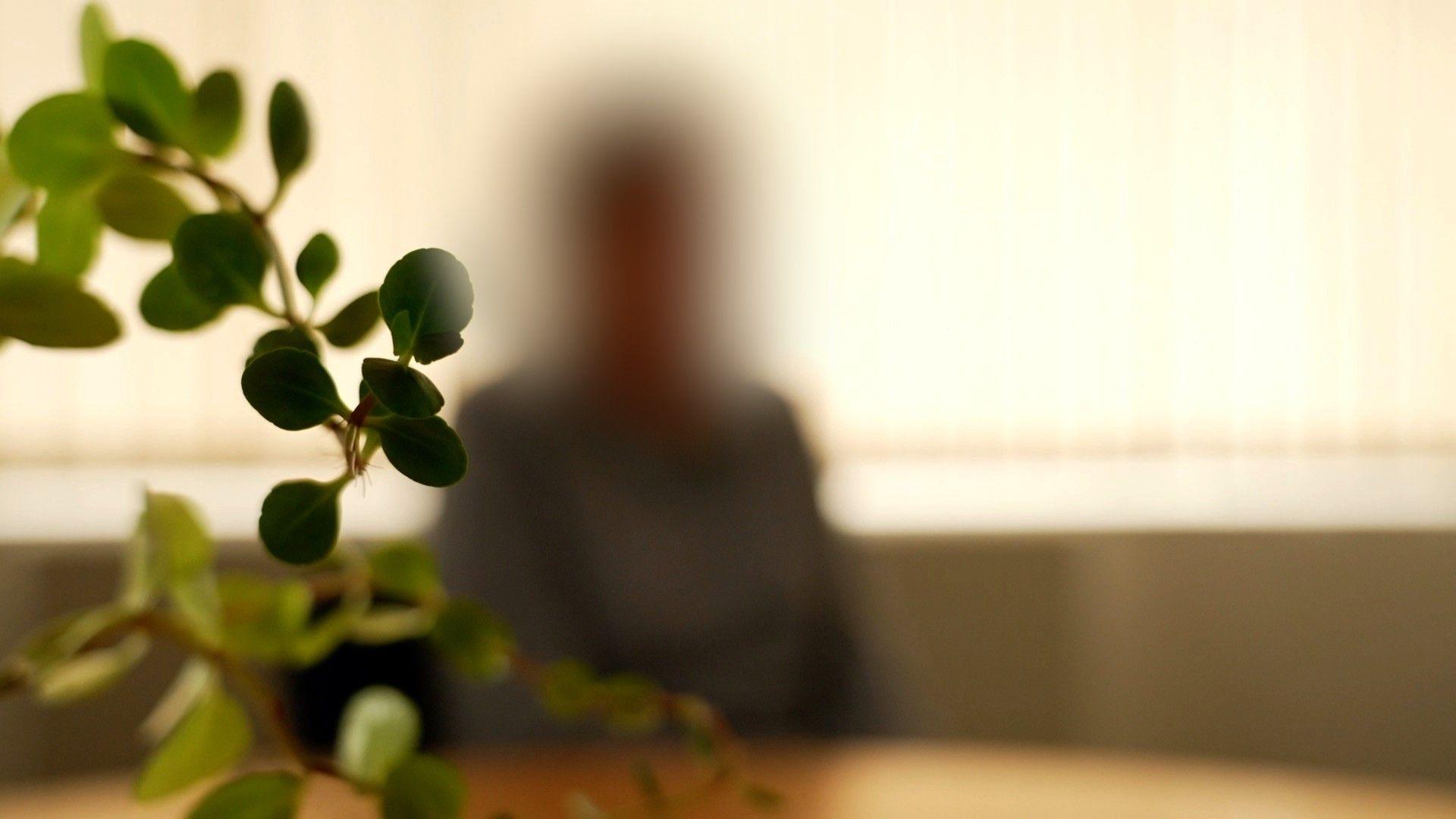 A blurred figure sits on a seat with a light-coloured table in the foregound, and the leaves of a pot plant.