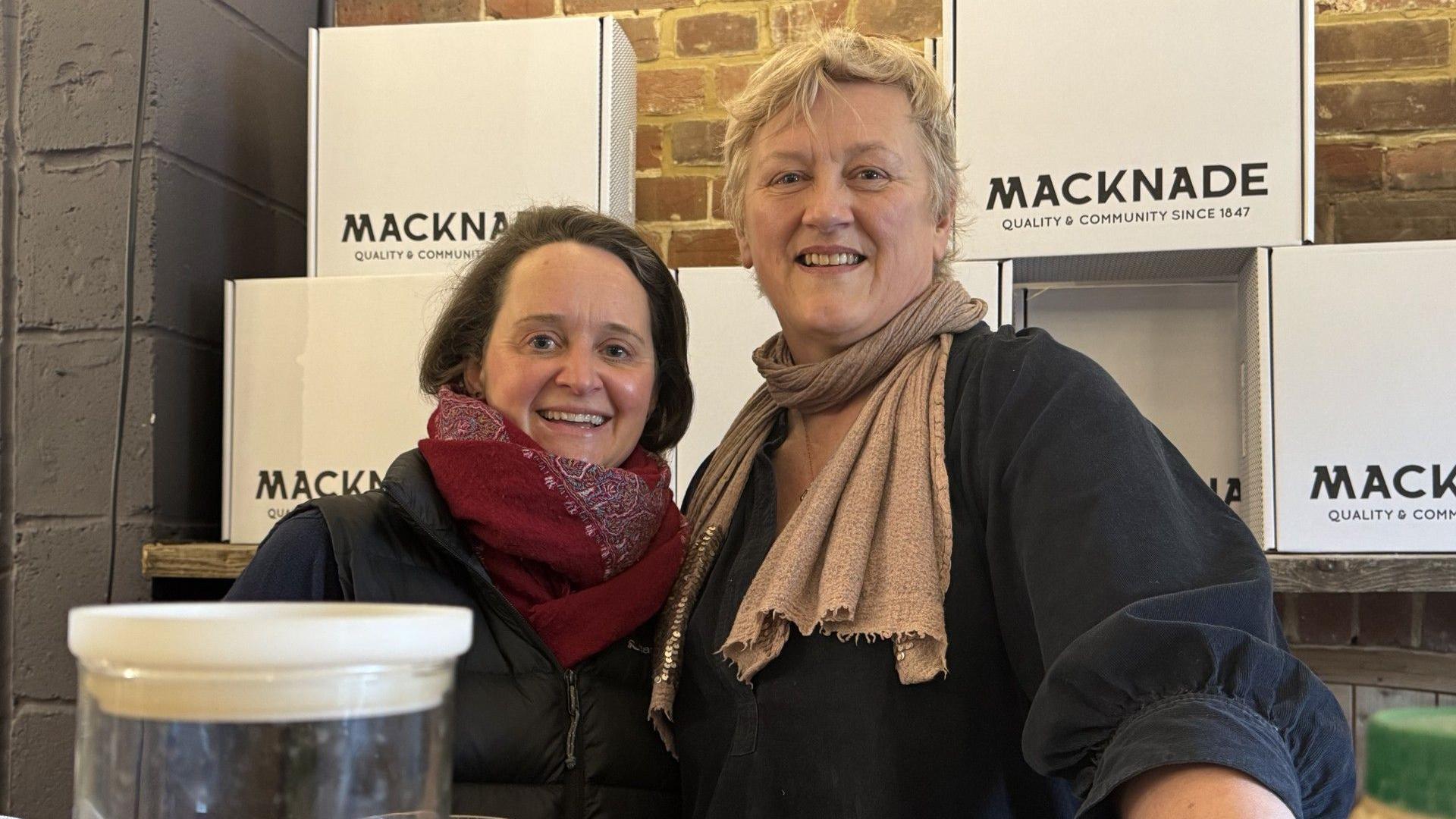 A woman with shoulder-length brown hair wearing a black coat and red scarf standing next to a blonde woman with short hair which is wearing a black shirt and beige scarf.