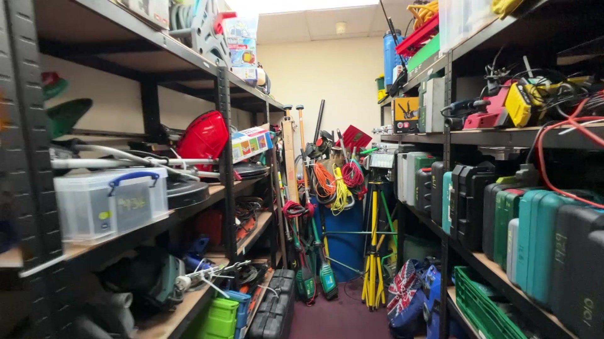 Stock at the Library of Stuff, with two rows of shelves on either side of the photo. The shelves are filled with various items, including toolboxes and tools. The carpet is purple and the walls are cream. The ceilings are tiled and white. 