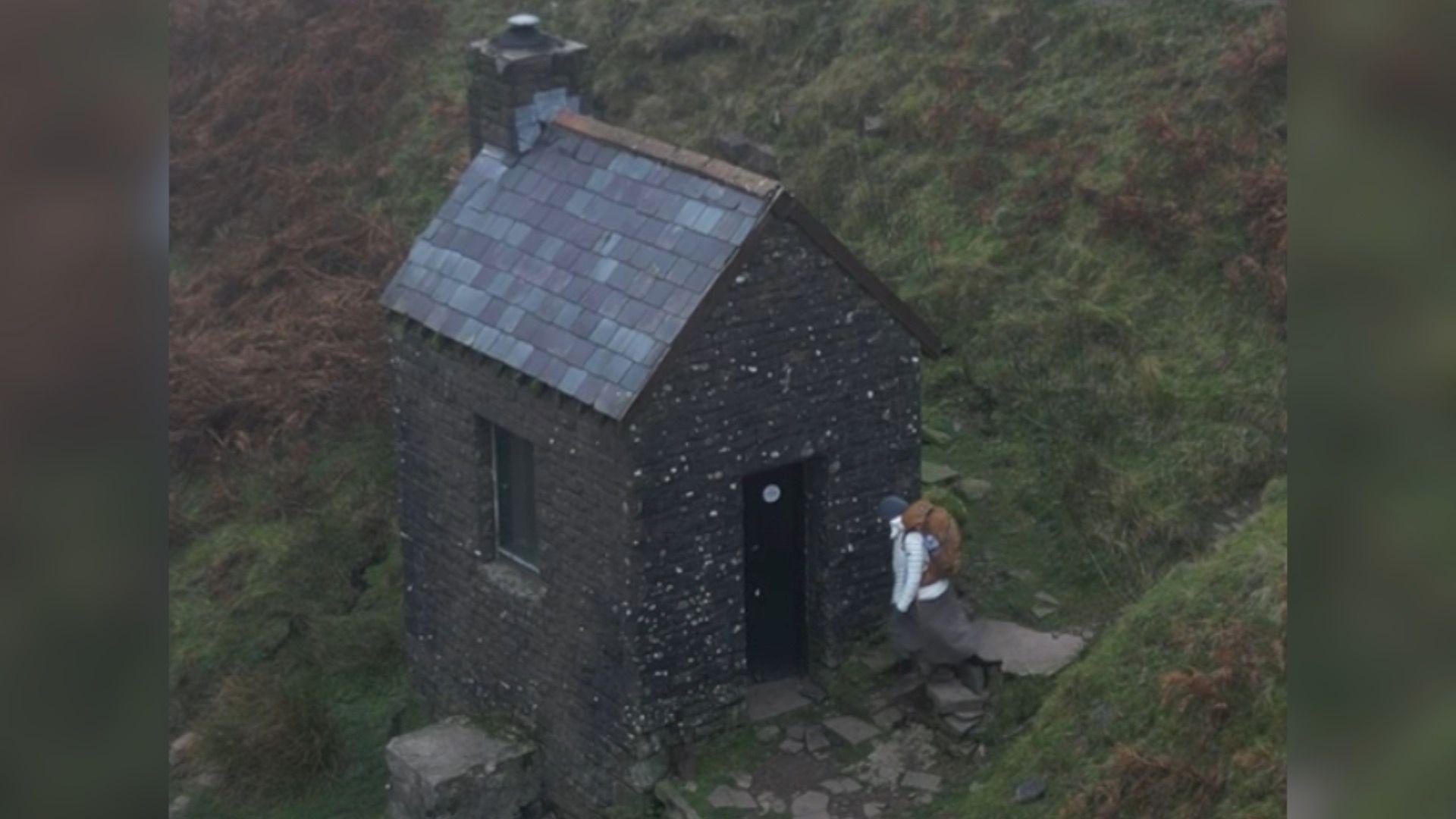 Amina approaching a stone bothy surrounded by countryside. The small building has a door on one side and a window on the other, with a slate roof and a chimney. 