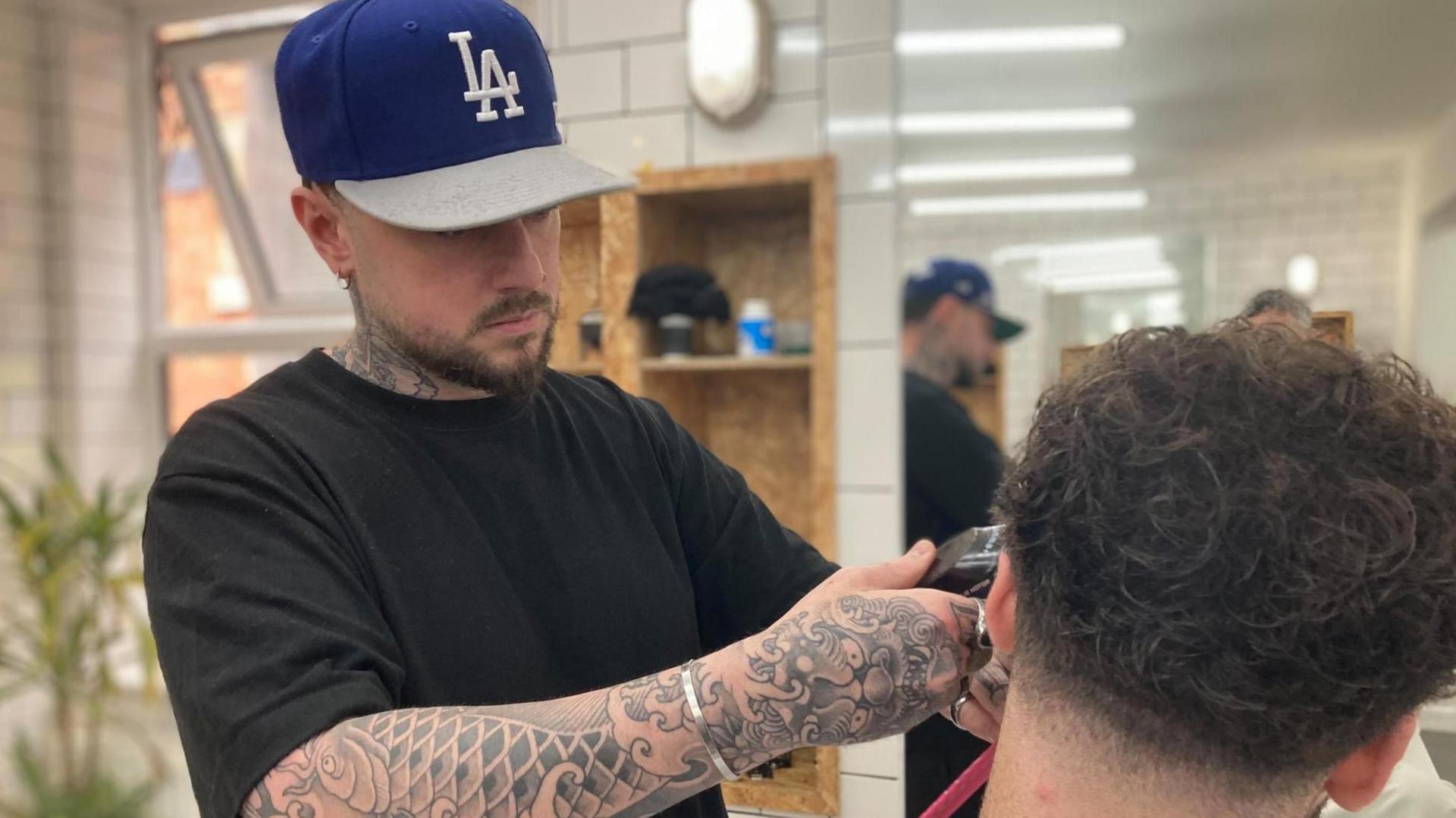 A young man with a baseball cap and tattoos on his arms holds an electric razor up to a mirror