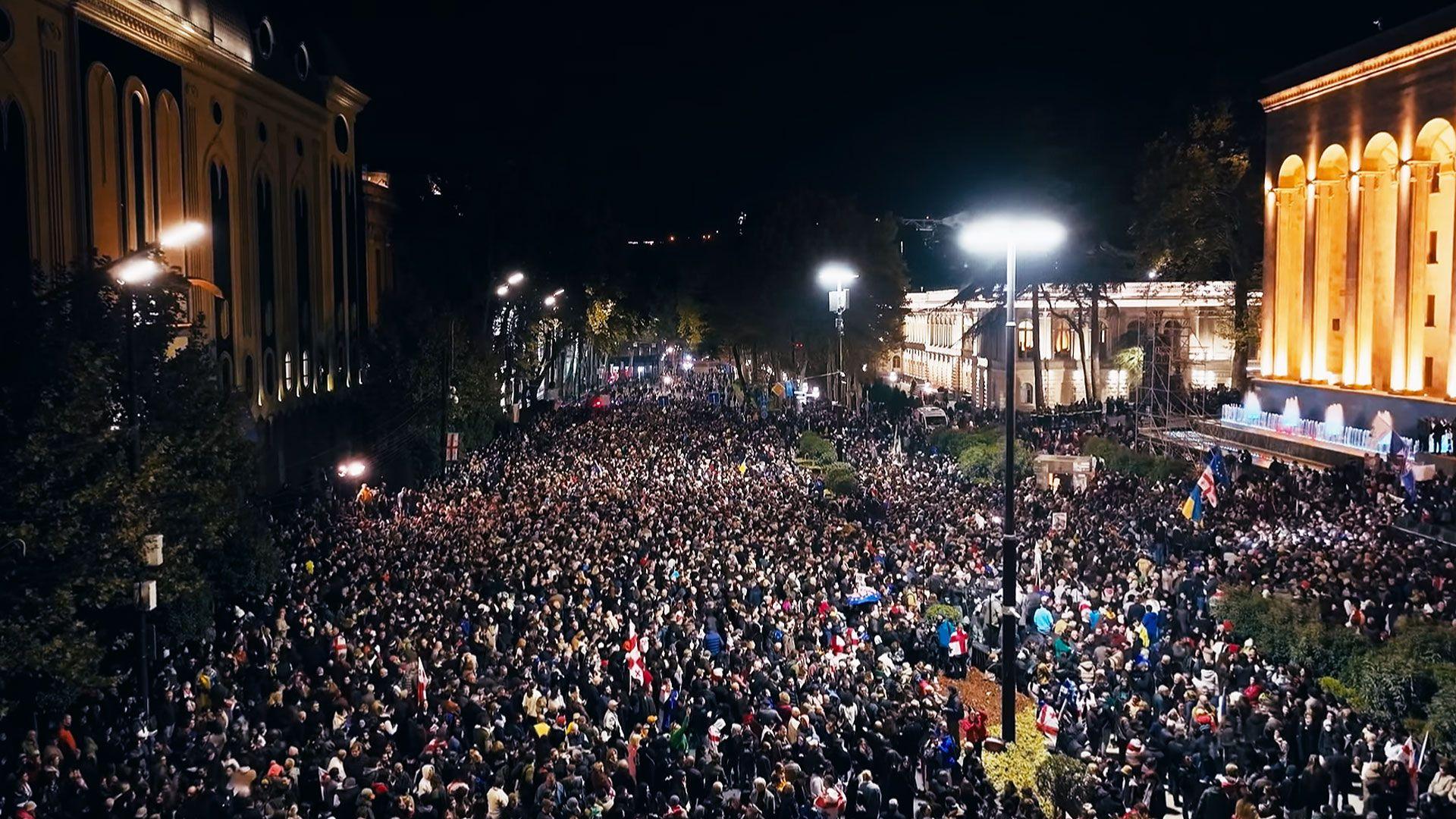 Hundreds of people demonstrating in the Georgian capital - massing on the street in the darkness with the parliament lit up to the side. Some EU flags are visible in the crowd
