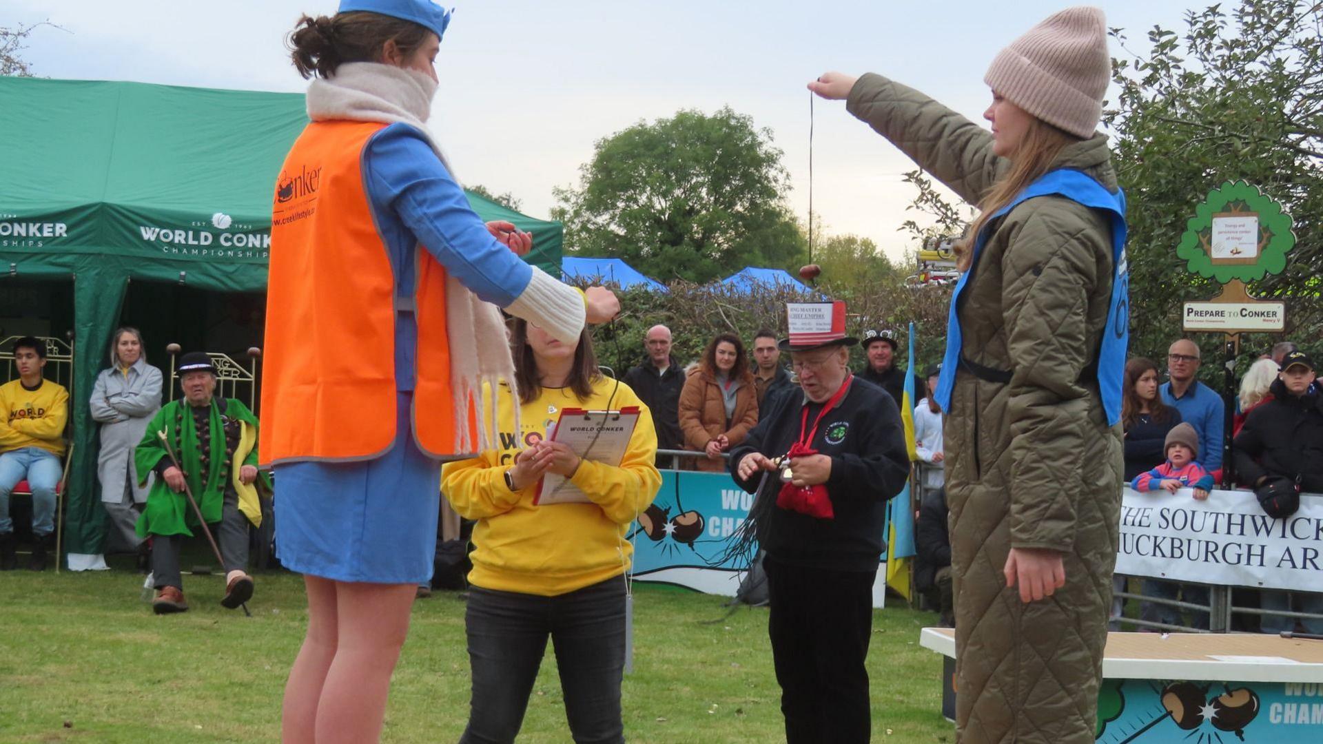 On the left, a woman dressed as an air hostess prepares to play conkers with Kelci, who is wearing a wool hat, and is standing on the right and dangling her conker on a string at shoulder height. A crowd of spectators is gathered around them.