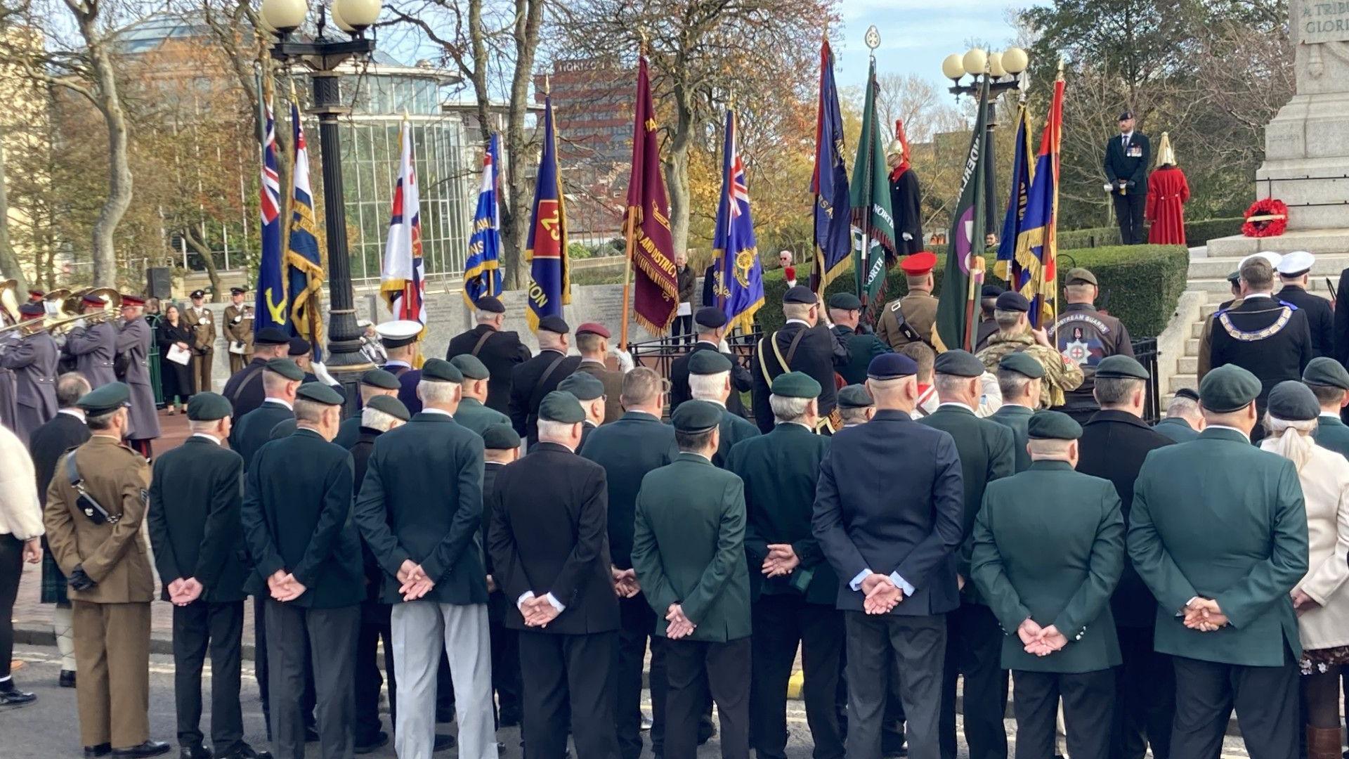 Armed forces veterans stand at ease in front of Burdon Road cenotaph in Sunderland. A poppy wreath has been laid at the base of the monument. A number of flags have been raised.