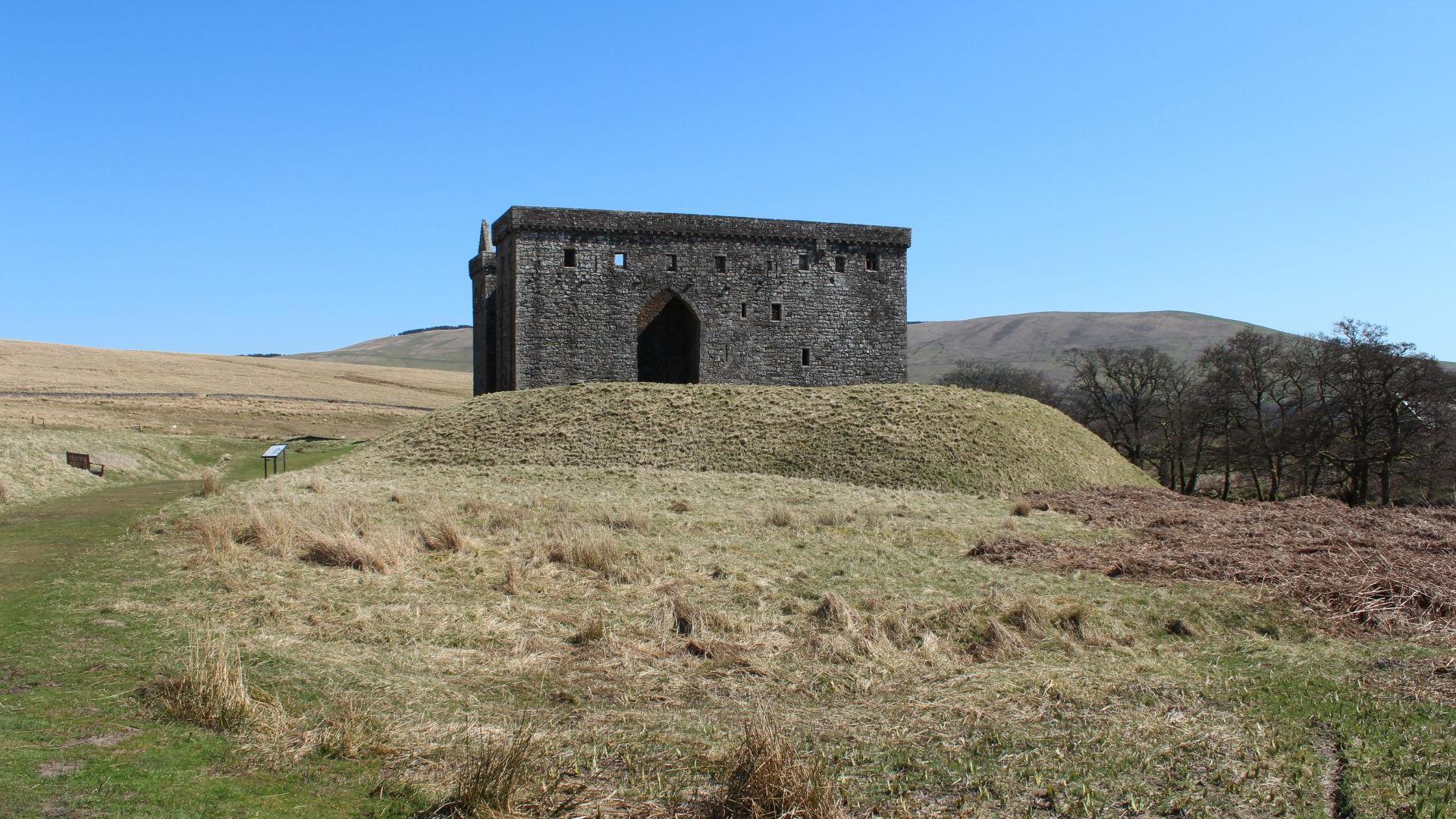 The remains of an ruined castle sit on top of a mound of earth in the rolling Borders landscape beneath a blue sky