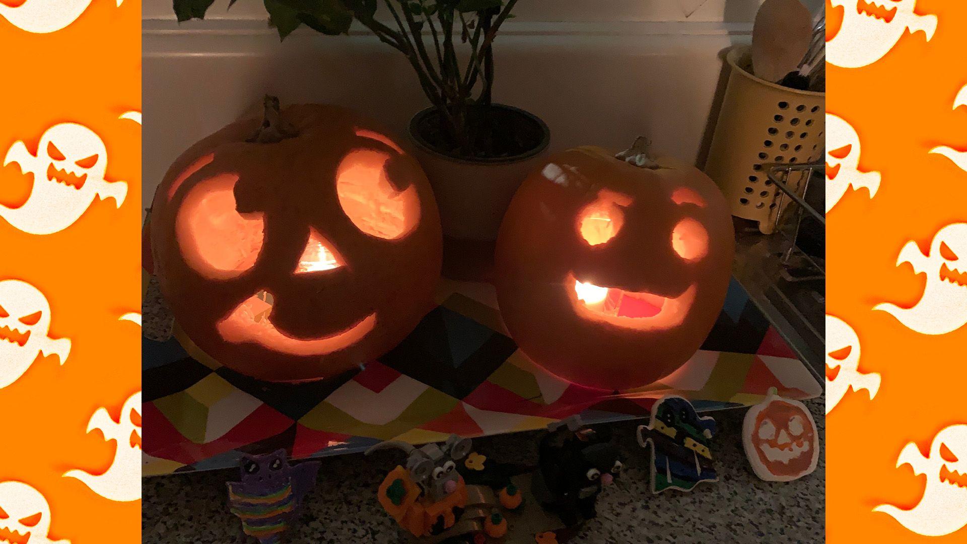 Two pumpkins have been carved and have a candle lit on the inside showing their designs. The pumpkin on the left has a triangular nose, a smile slanting to the left and two eyes looking to the right with its eyebrows raised. On the right, the pumpkin has a wide open-mouthed smile, two eyes and one eyebrow raised