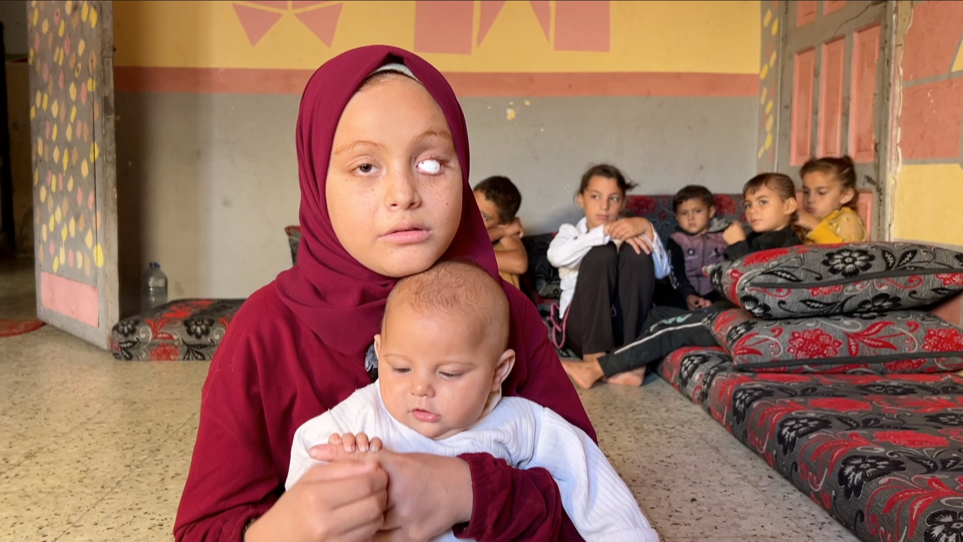 A young teenage girl - Malak - holds her baby sister, Rahma, on her lap. Malak is wearing a burgundy headscarf and long-sleeve tunic. One of her eyes has been replaced with a white sphere. She is looking to the side of the camera. The baby is holding onto her sister's finger and looking at it. She is wearing a long-sleeved white top. In the background, their five siblings sit close together in the corner of a bare room with its walls painted with square patterns,  on large floor cushions. 