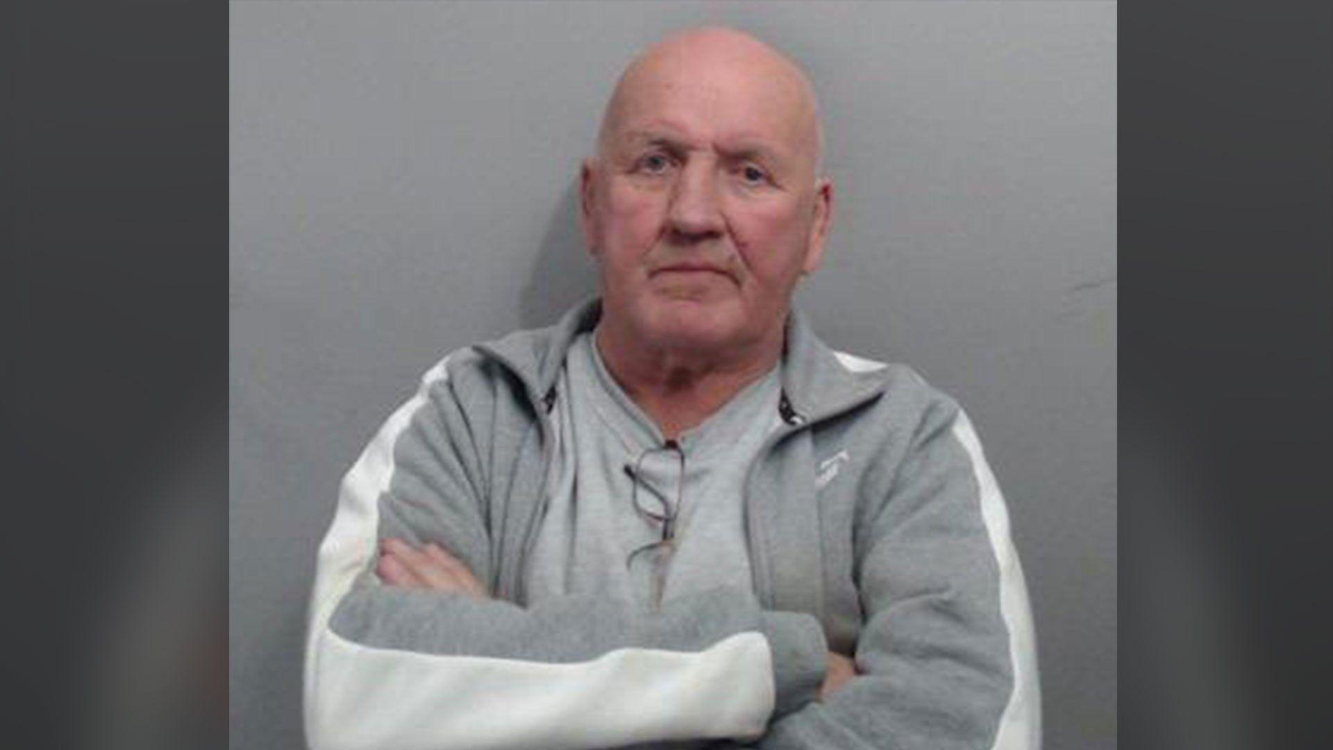 A man, standing in front of a grey background. He is bald and has his arms crossed. He is wearing a grey t-shirt and grey zip up jumper.